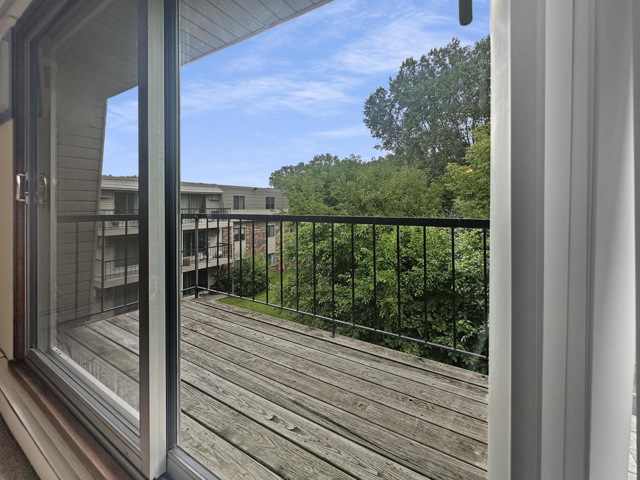 View from a window showing a balcony with a wooden floor. Outside, lush green trees and grassy areas are visible, along with the exterior of an apartment complex in the background. The sky is clear with a few clouds, creating a bright and inviting atmosphere.