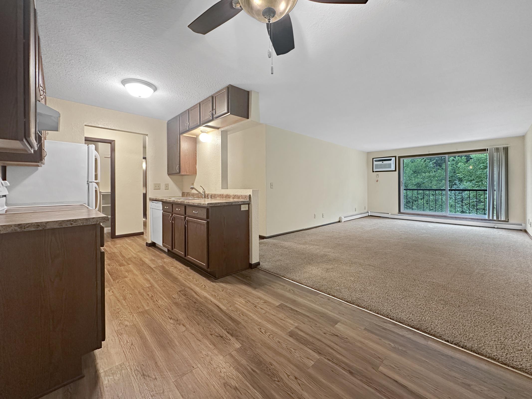 A spacious living area with large windows and carpet flooring, adjacent to a kitchen featuring dark wooden cabinets and white appliances. Bright overhead lighting and a ceiling fan are present. The room offers a door leading to an outdoor space, enhancing the natural light.