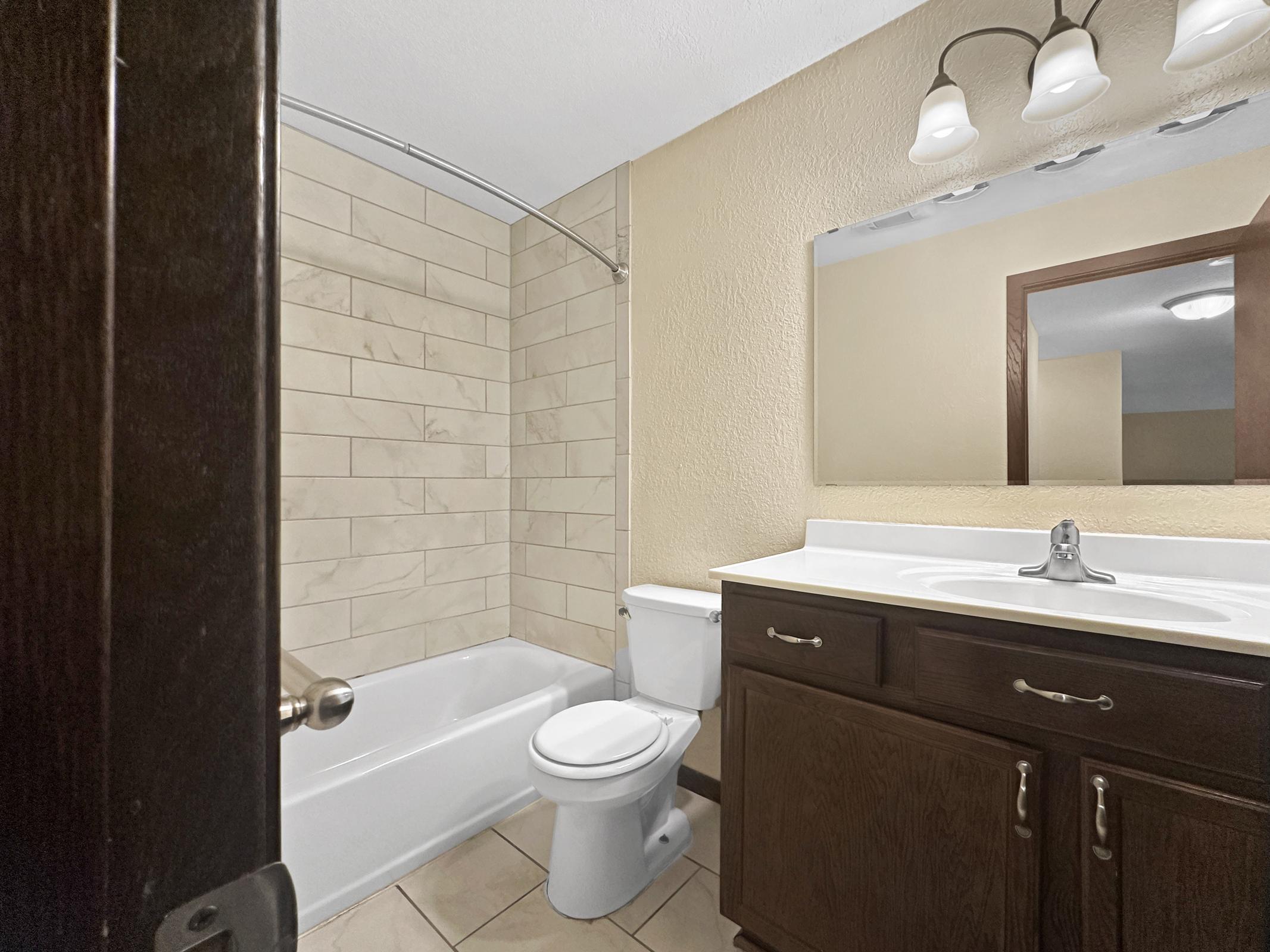 A clean, modern bathroom featuring a beige tiled shower and bathtub combination, a white toilet, and a dark wood vanity with a sink. The wall is painted in a light beige color, and there's a large mirror above the vanity. Soft overhead lighting illuminates the space.