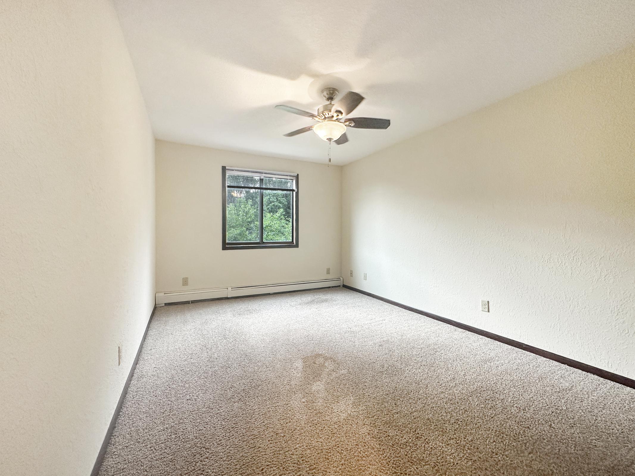 A spacious, empty room with beige wall color and carpeted flooring. A ceiling fan is mounted in the center, and a large window on one wall allows natural light in, revealing greenery outside. The room has a simple and clean design, ideal for personal decoration or furnishing.