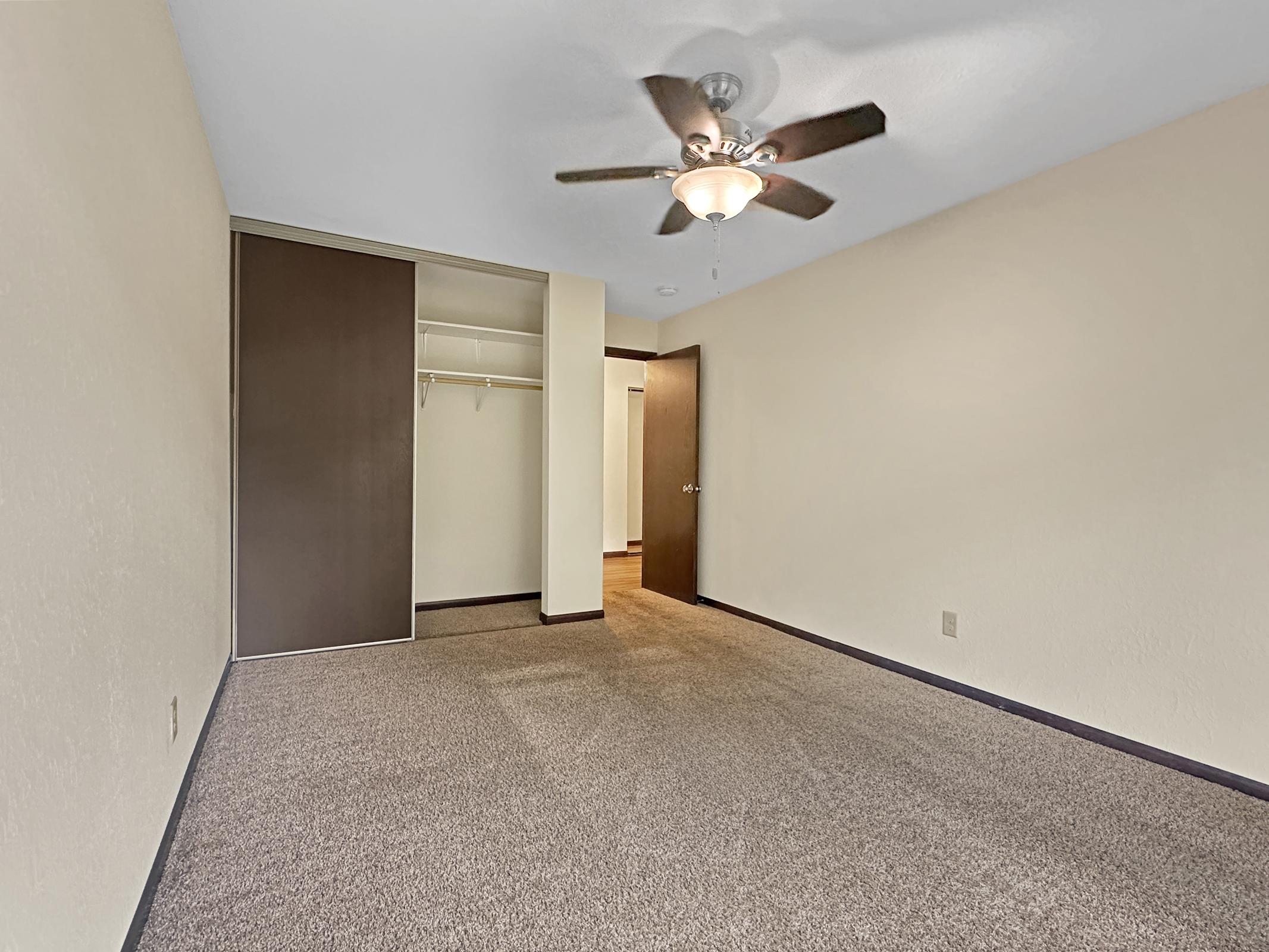 A spacious, unfurnished room with light beige walls and carpeted flooring. A ceiling fan is installed, and there’s a closet with sliding doors on one side. A doorway leads to another area of the room, creating an open and airy atmosphere. Natural light appears to be entering the space.