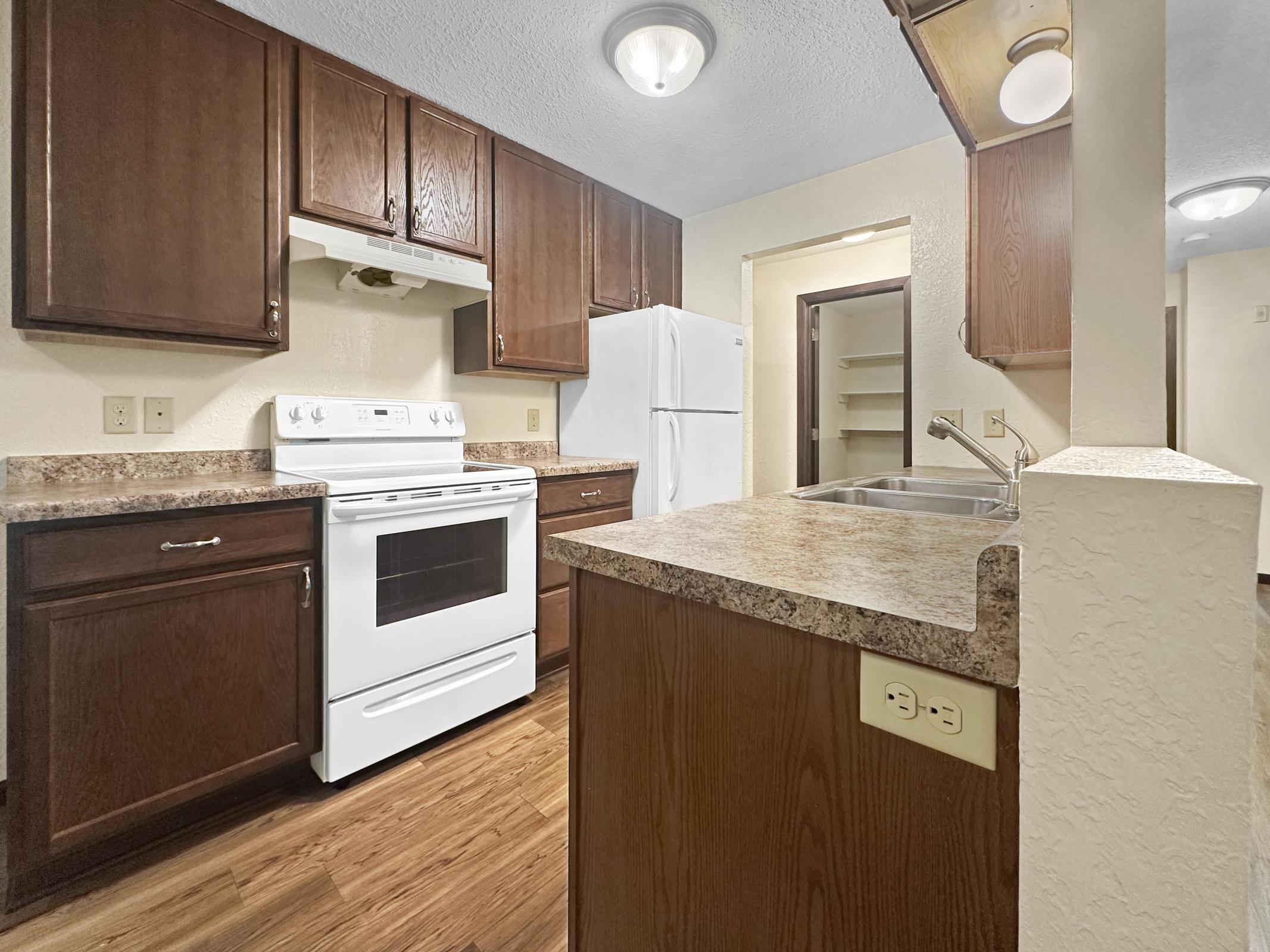 Modern kitchen featuring wooden cabinets, a white stove and refrigerator, and a dual basin sink. The countertops are brown and textured, and there's overhead lighting. A pantry door is visible in the background, highlighting a functional and inviting cooking space.