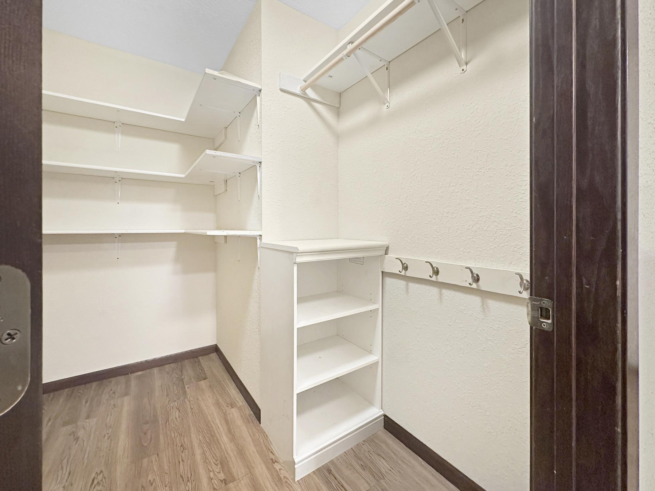 Empty closet with white shelves and a small built-in shelf unit. The walls are a light color and the floor has brown wooden laminate. There are hooks mounted on the side for hanging items, and the door is partially visible.