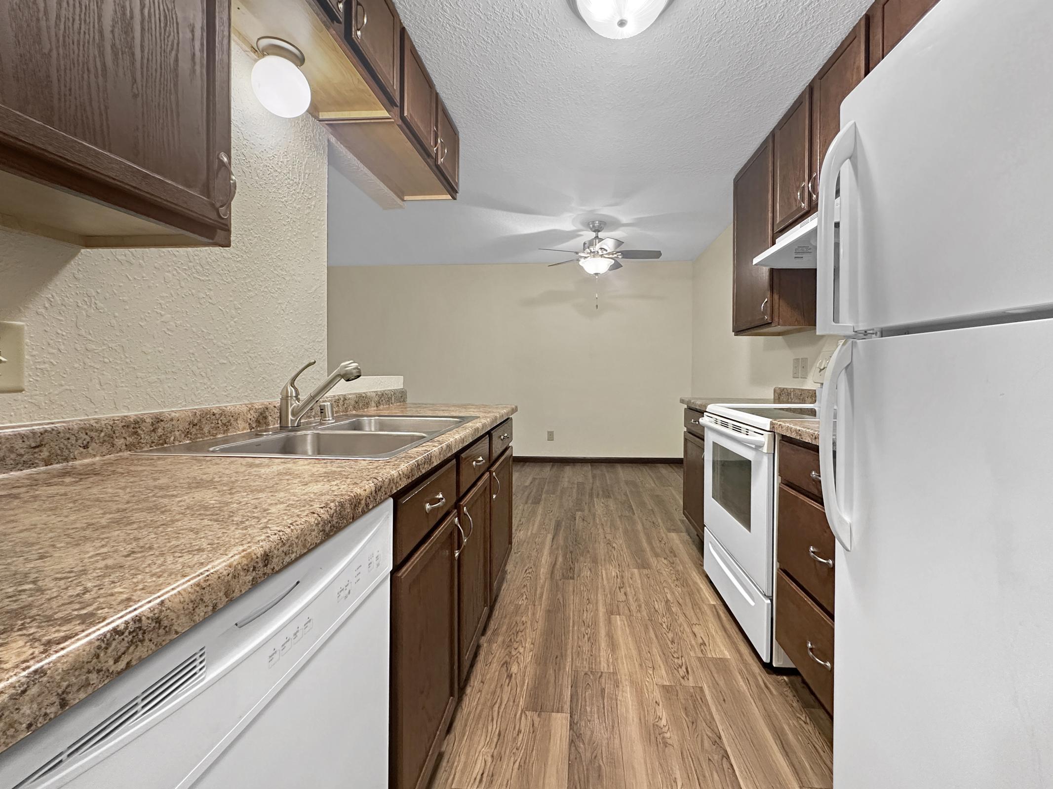 A clean and modern kitchen featuring dark wooden cabinets, a double sink, and granite countertops. There is a white refrigerator and stove, with a ceiling fan visible. The floor is covered in laminate wood, and the background shows a well-lit, open space.