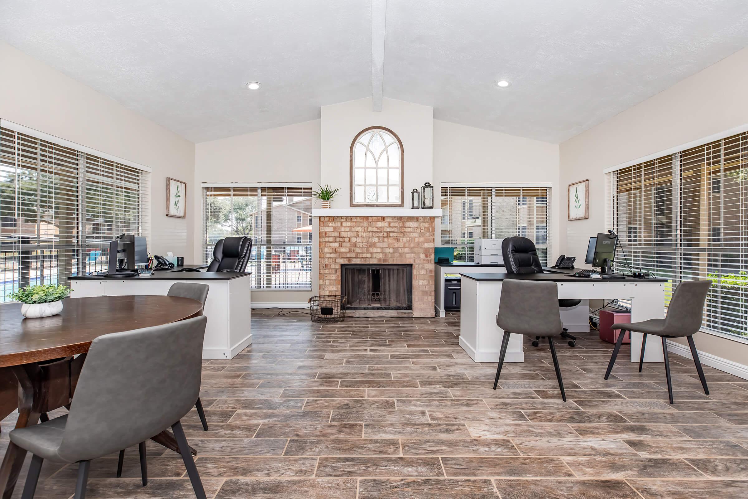 Bright and spacious office area featuring two desks with computers, a dining table, and a brick fireplace. Large windows allow natural light, and contemporary furnishings create a modern atmosphere. The warm tones and neutral decor enhance the inviting ambiance of the workspace.
