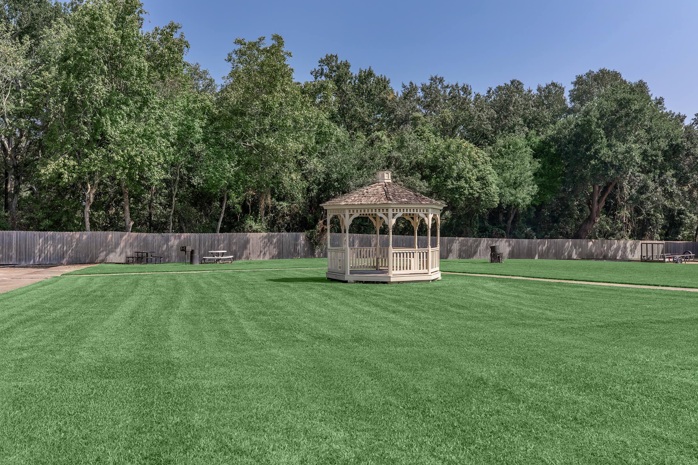 A well-manicured green lawn featuring a white gazebo surrounded by dense trees and a clear blue sky. Benches and picnic tables are visible in the background, providing a tranquil outdoor setting.