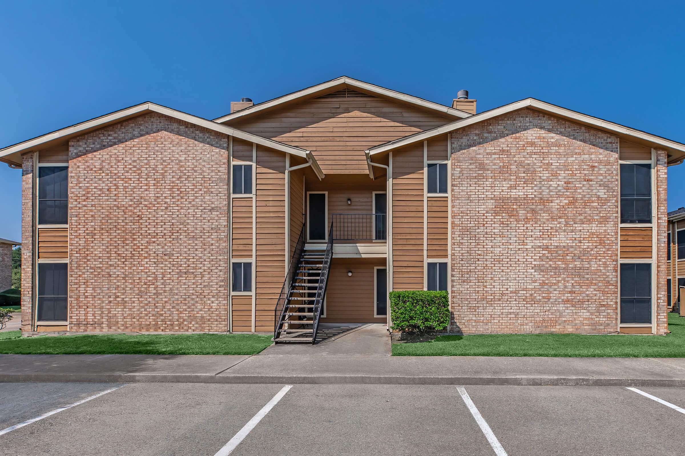 A two-story brick apartment building with a symmetrical façade, featuring two separate entrances with wooden stairs leading to the second floor. The building has large windows and green grass in front, with a paved parking lot in the foreground. The sky is clear and blue.