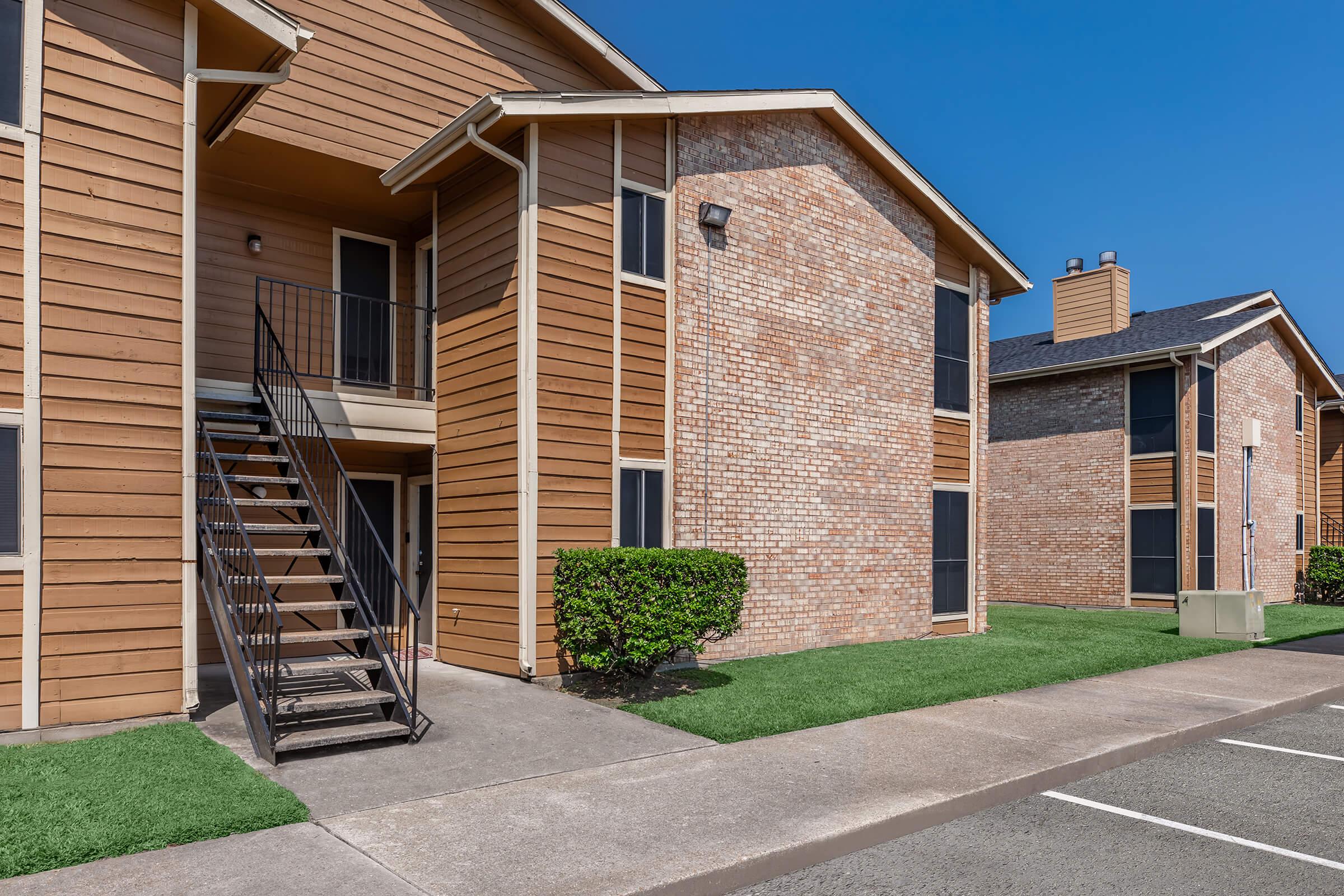 A view of a multi-unit apartment building featuring two stories, wooden siding, and brick accents. A set of stairs leads to a second-floor entrance. The scene includes neatly trimmed grass and a sidewalk, with clear blue skies in the background.