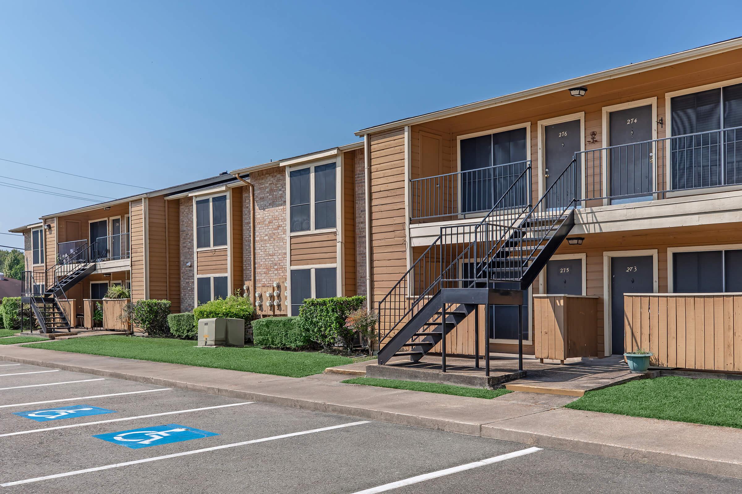 A row of two-story apartment buildings with wooden exteriors. Each unit has a stairway leading to the entrance. The foreground features designated parking spaces with blue accessibility symbols. Well-maintained grass and small shrubs line the walkway between the buildings. Bright blue sky in the background.