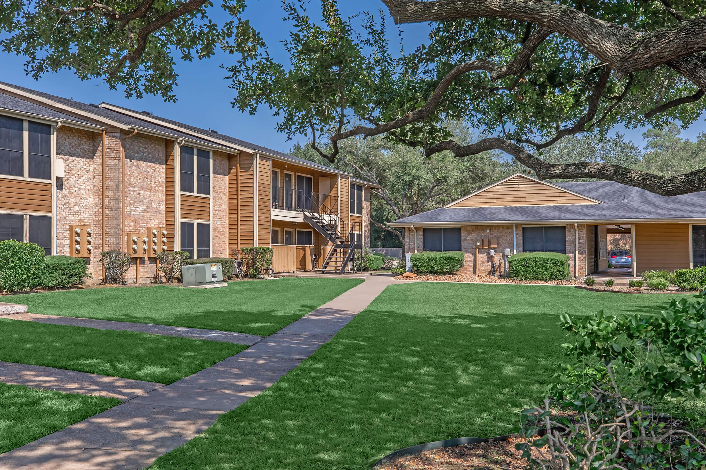 A well-maintained apartment complex surrounded by green lawns and trees. The buildings feature a mix of brick and wood siding, with balconies on the upper floors. A walking path runs through the grounds, leading to a centrally located community building. The sky is clear and sunny, enhancing the inviting atmosphere.