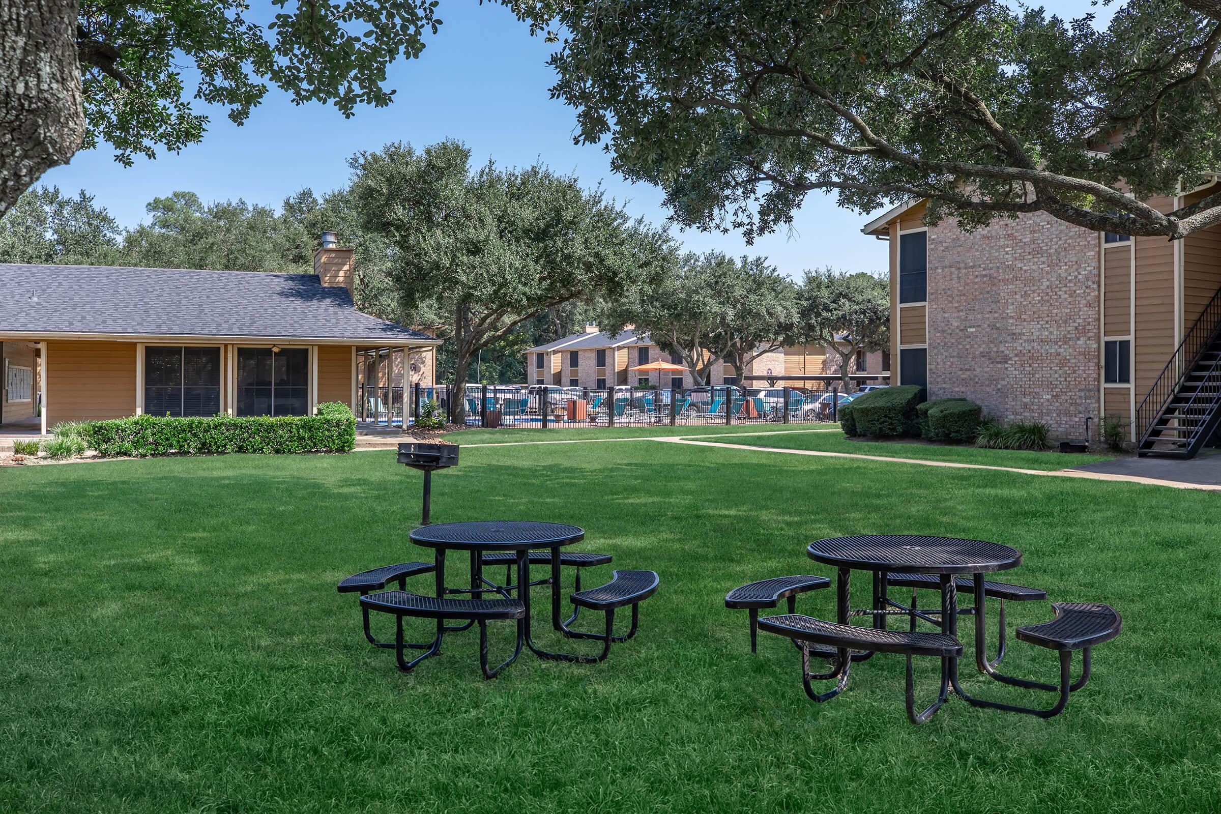 A grassy area featuring a black picnic table with attached benches, surrounded by trees. In the background, there are two buildings, one with brick and the other with a yellow exterior. A pool area is visible beyond the fence, indicating a residential complex with outdoor communal spaces.