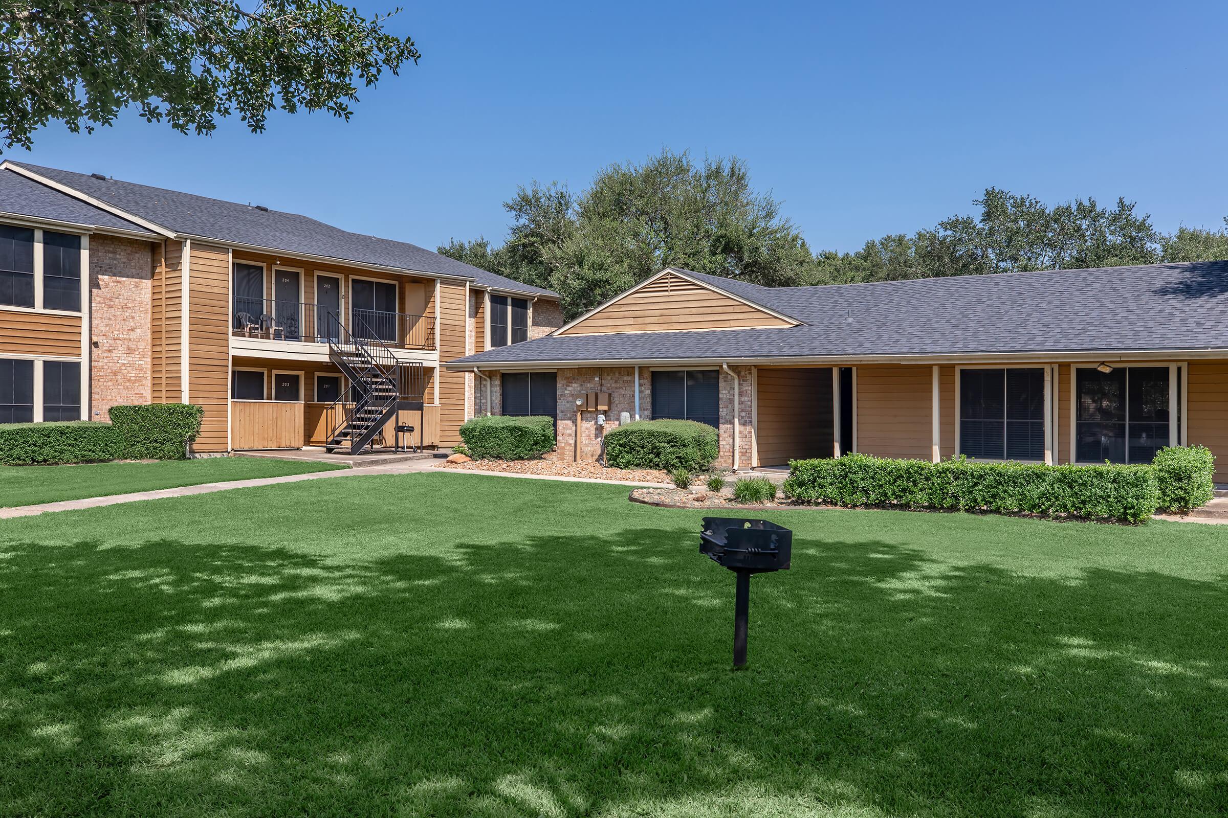A well-maintained apartment complex featuring two buildings surrounded by lush green lawns. The structures have brown siding and a modern design, with a staircase leading to upper units. A small, black grill is situated on the grassy area in the foreground. Clear blue skies above create a welcoming atmosphere.