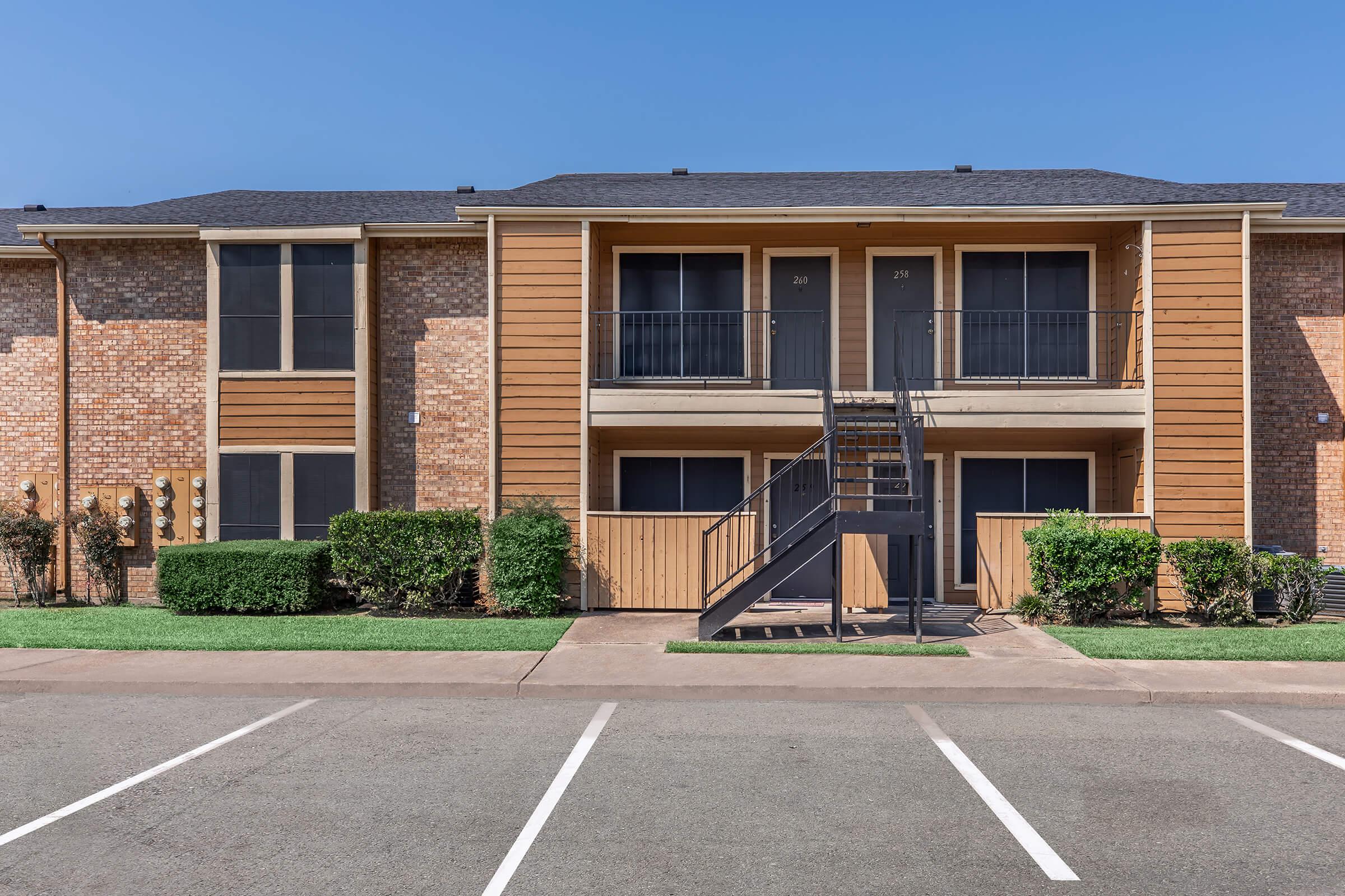 Exterior view of a residential apartment complex featuring two stories, wooden balconies, and a black metal staircase. The building has a mix of brick and wood siding with green lawn and shrubs in front. Several parking spaces are visible in the foreground under a clear blue sky.