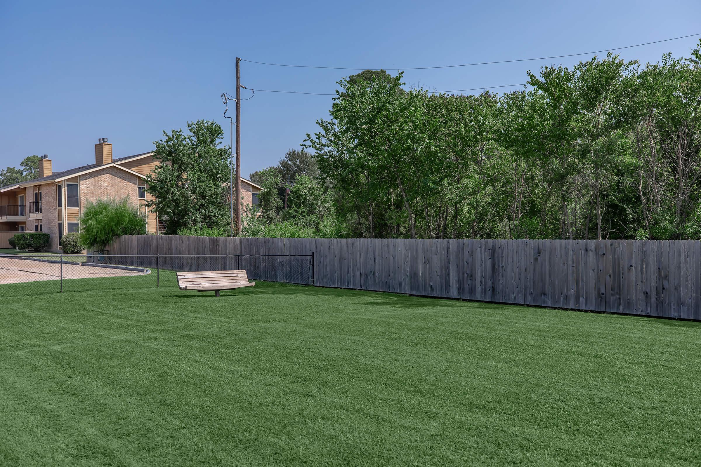 A spacious green lawn with synthetic turf, bordered by a wooden fence. In the background, there are trees and a residential building partially visible. A simple wooden bench sits on the lawn, providing a place to relax. The sky is clear and blue, indicating a bright day.