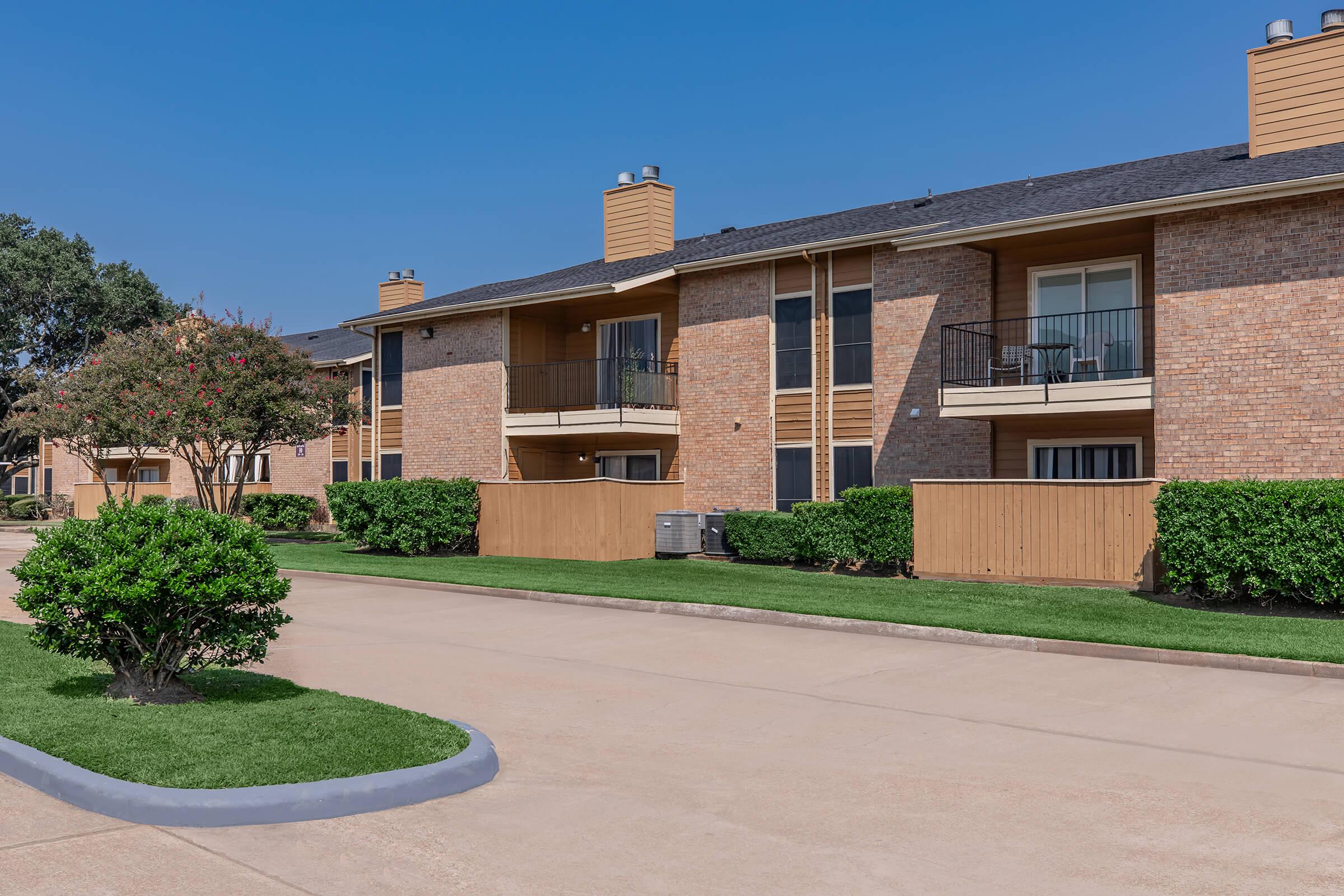 Two-story apartment complex featuring brick exteriors and wooden balconies. Neatly landscaped with green shrubs and grass lining the walkway. Clear blue sky overhead and a paved road in front.