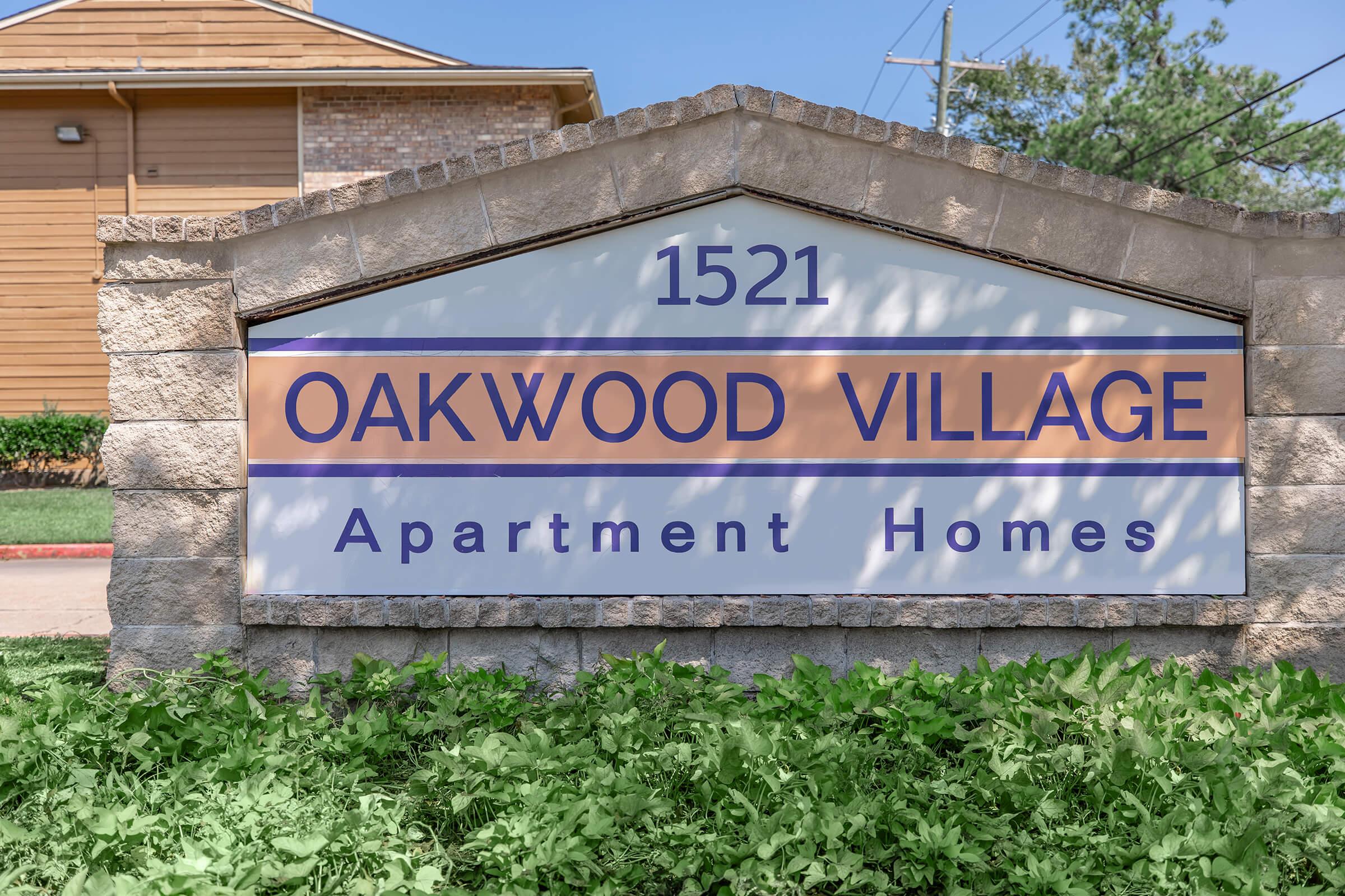 Sign for Oakwood Village Apartment Homes displaying the address "1521" prominently. The sign is stone-carved with a white background and orange and purple text. Surrounding greenery adds a natural touch, and the setting appears sunny and welcoming.