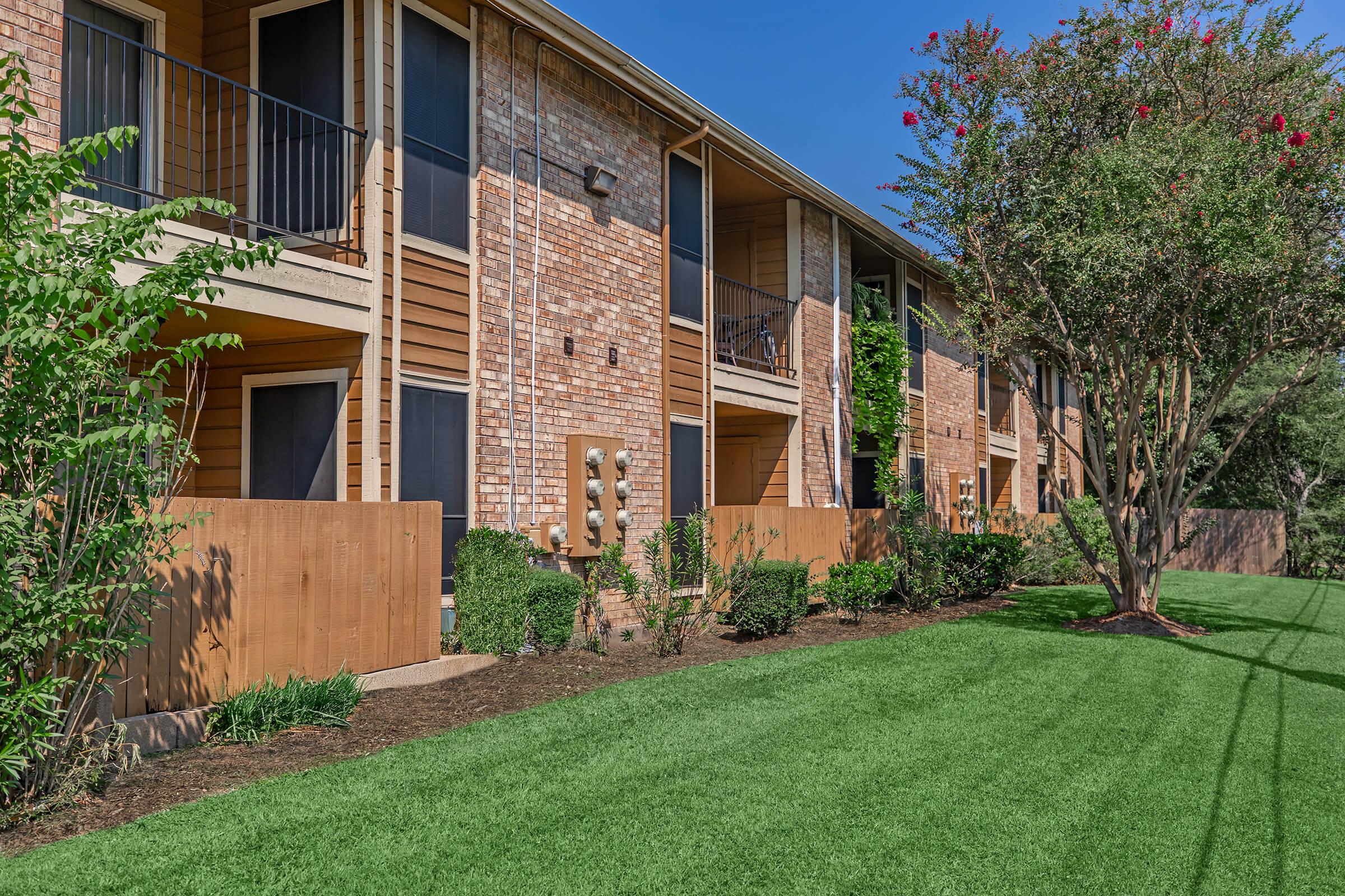 A well-maintained multi-unit apartment building with wooden balconies and brick exterior. The surrounding area features lush green grass and neatly trimmed bushes, along with wooden fences and a tree in bloom, set against a clear blue sky.