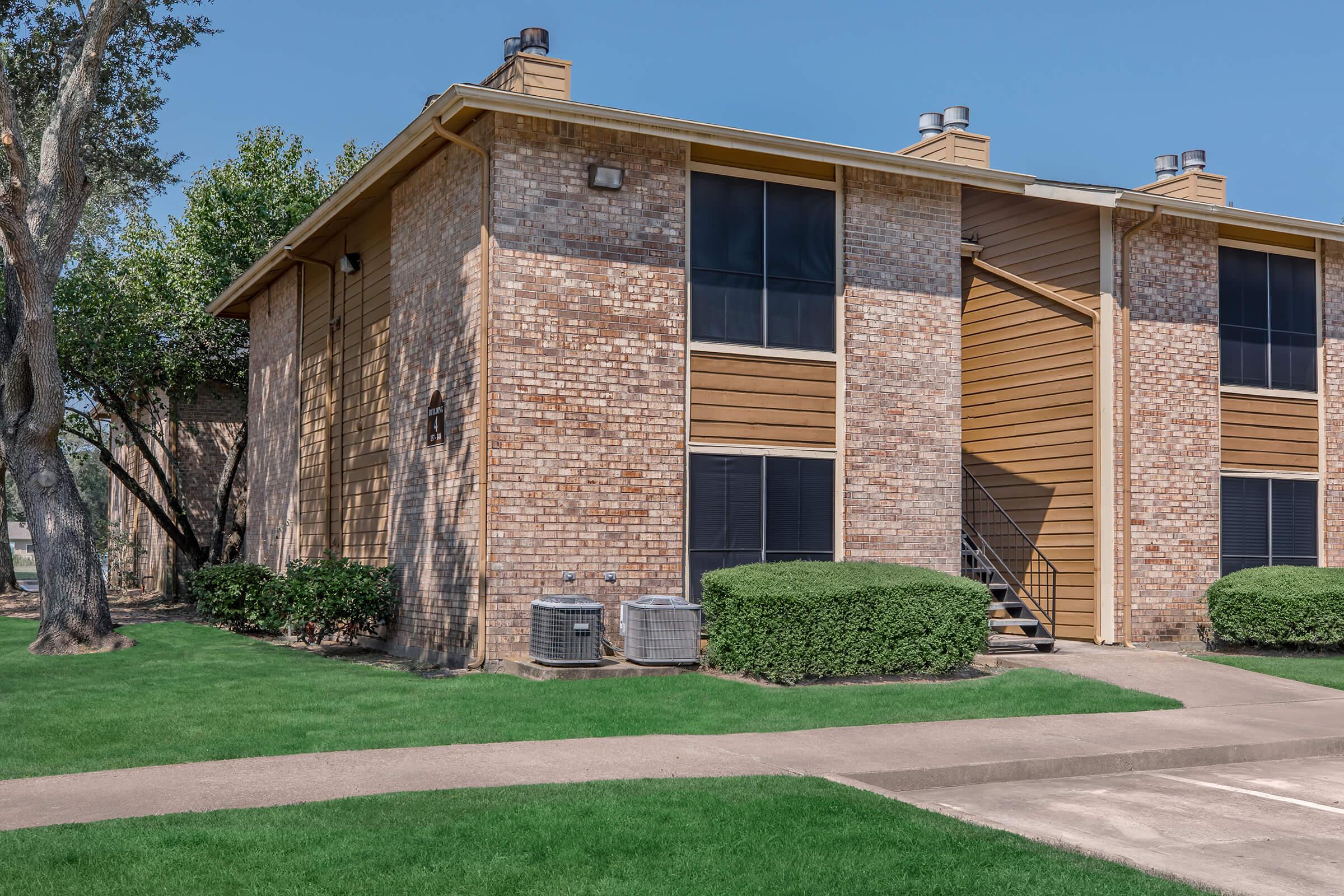 Two-story brick apartment building with wooden accents, featuring large windows and two outdoor air conditioning units. The building is surrounded by green grass and neatly trimmed bushes, with a concrete walkway leading up to the entrance. Nearby trees provide some shade.