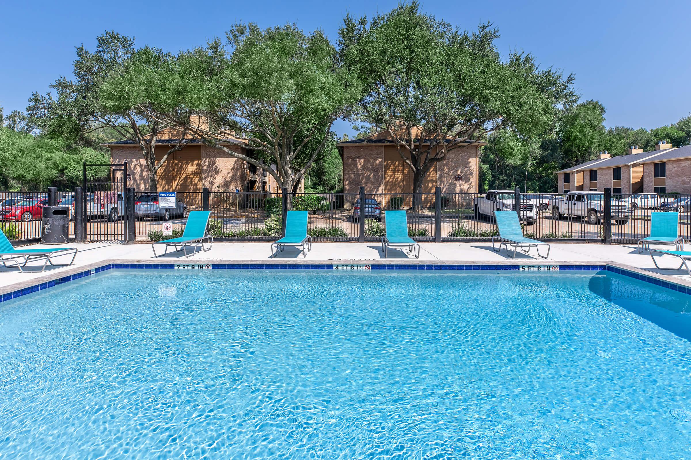 A clear swimming pool surrounded by lounge chairs, with trees in the background and residential buildings. The scene features a bright blue sky, creating a sunny and inviting atmosphere.