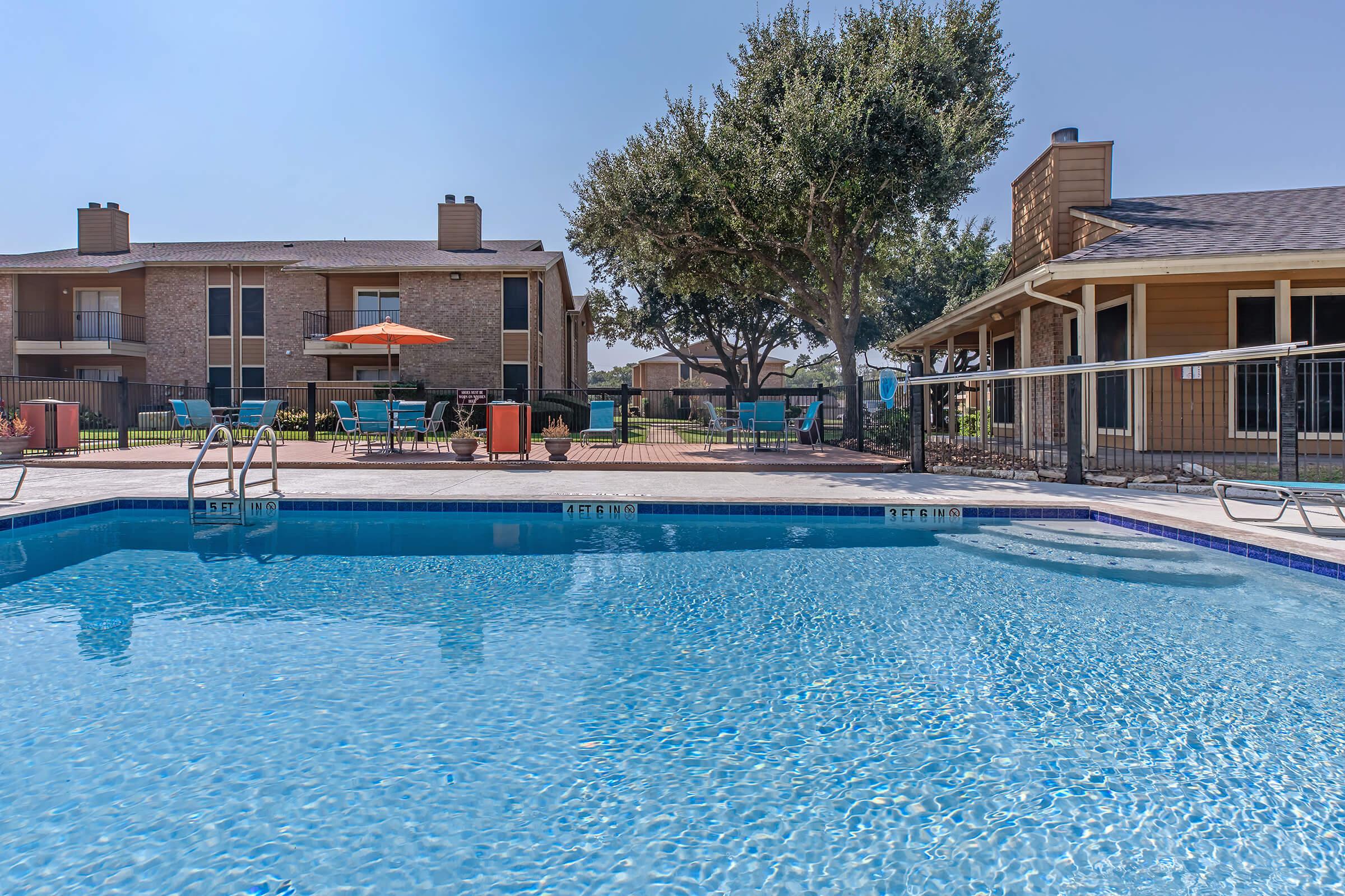 A clear blue swimming pool in the foreground, with lounge chairs and umbrellas around it. In the background, there are residential buildings and a clubhouse, surrounded by trees under a bright blue sky.