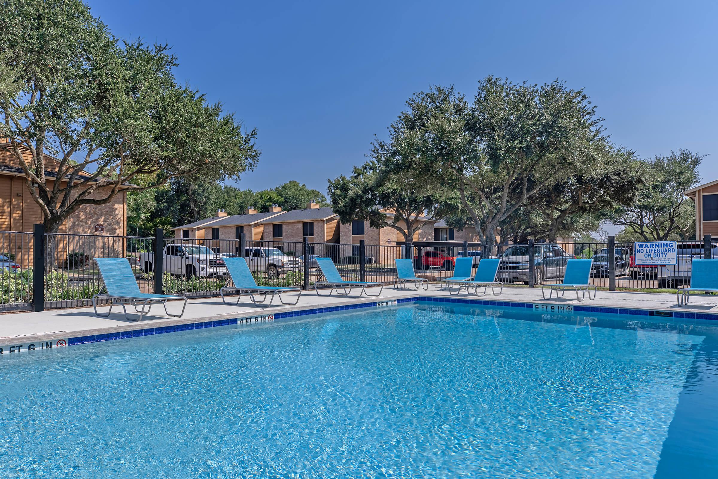A clear swimming pool surrounded by light blue lounge chairs under a bright blue sky. In the background, several residential buildings and trees are visible, along with parked cars and a warning sign near the pool area. The scene conveys a relaxing outdoor ambiance.