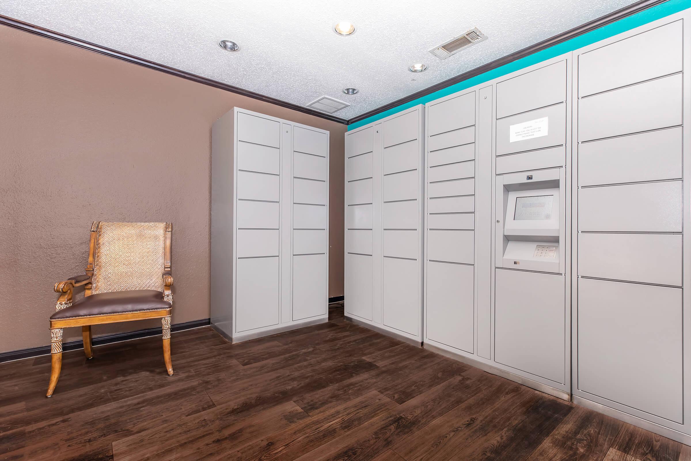 Interior view of a mailroom featuring multiple gray mail lockers, a small ATM, and a wooden chair. The walls are painted brown, and there is a strip of blue lighting along the top. The floor is wooden, giving a modern yet functional feel to the space.