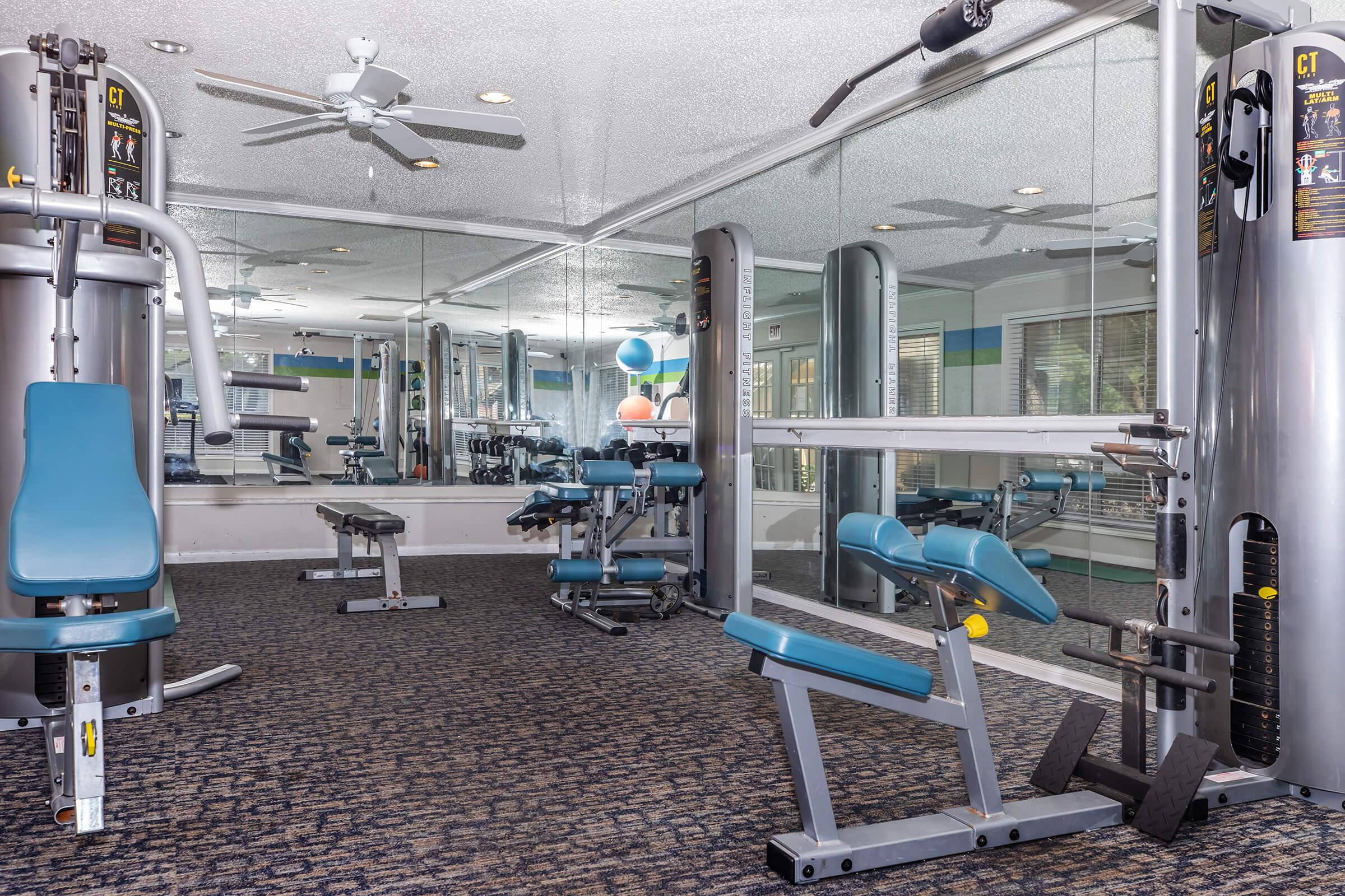 A well-equipped gym with workout machines, including a cable machine and bench. The room features large mirrors on the walls, a ceiling fan, and is carpeted. Brightly colored exercise balls are visible in the background, adding a vibrant touch to the fitness space.