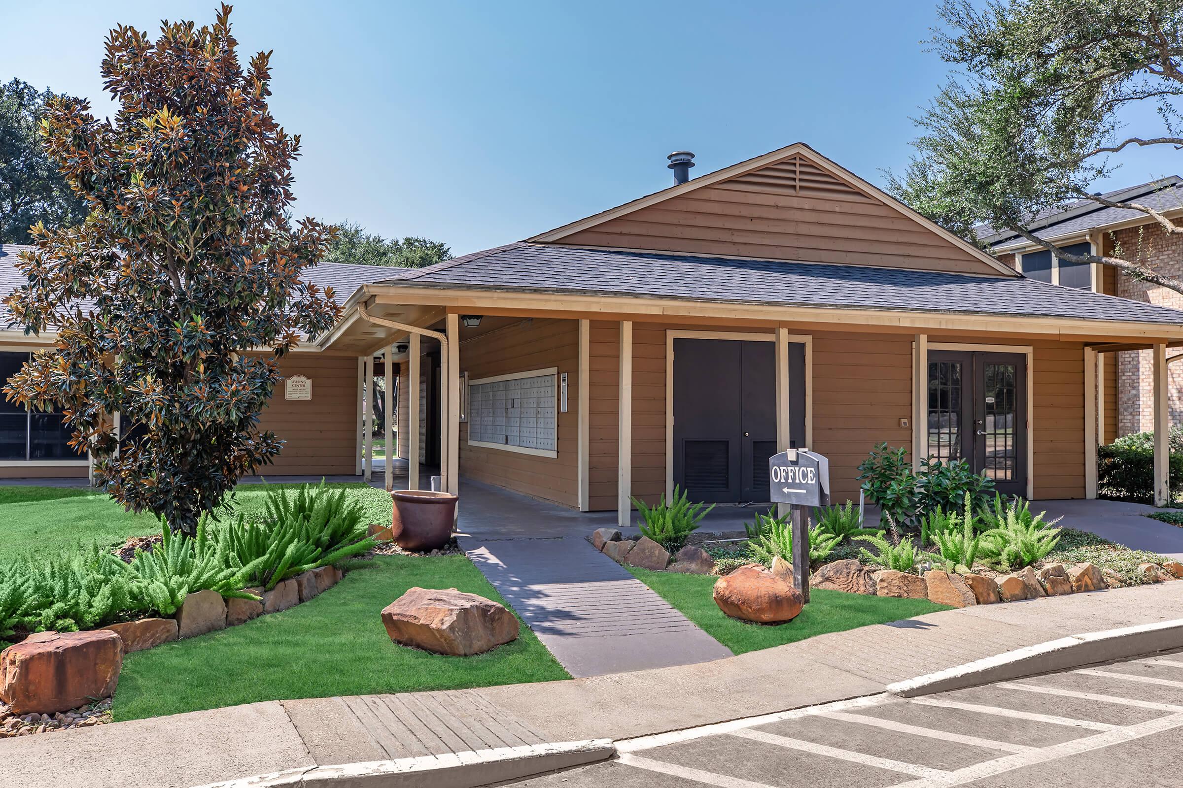 A modern office building with a light brown exterior, surrounded by landscaped greenery and rocks. The entrance features a sign labeled "Office," and there are neatly trimmed plants and trees in the foreground, adding to the inviting atmosphere.