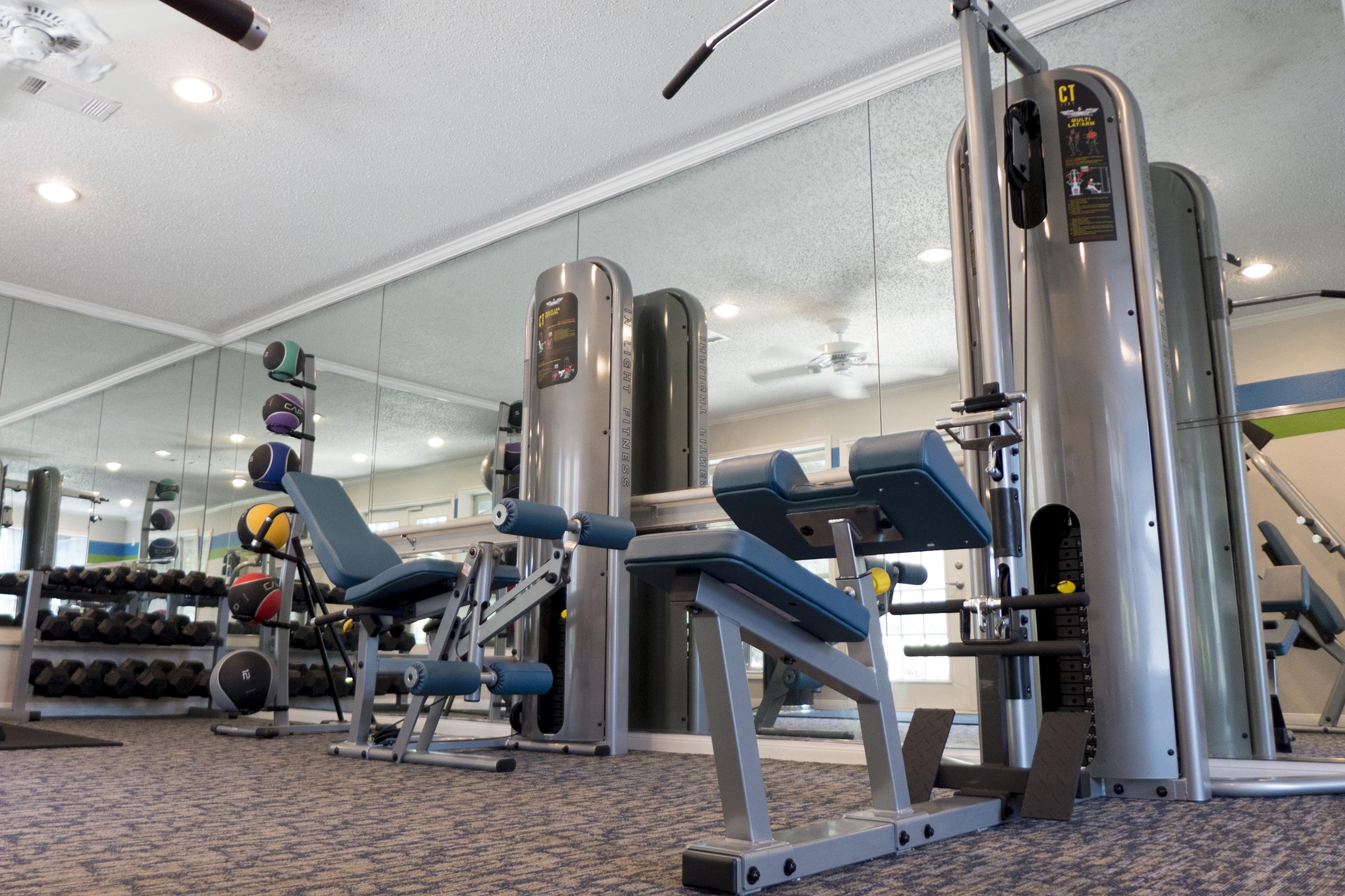 A well-equipped gym area featuring various workout machines, weights, and exercise balls. The space is bright with mirrors on the walls, allowing for an open and inviting atmosphere for fitness activities.