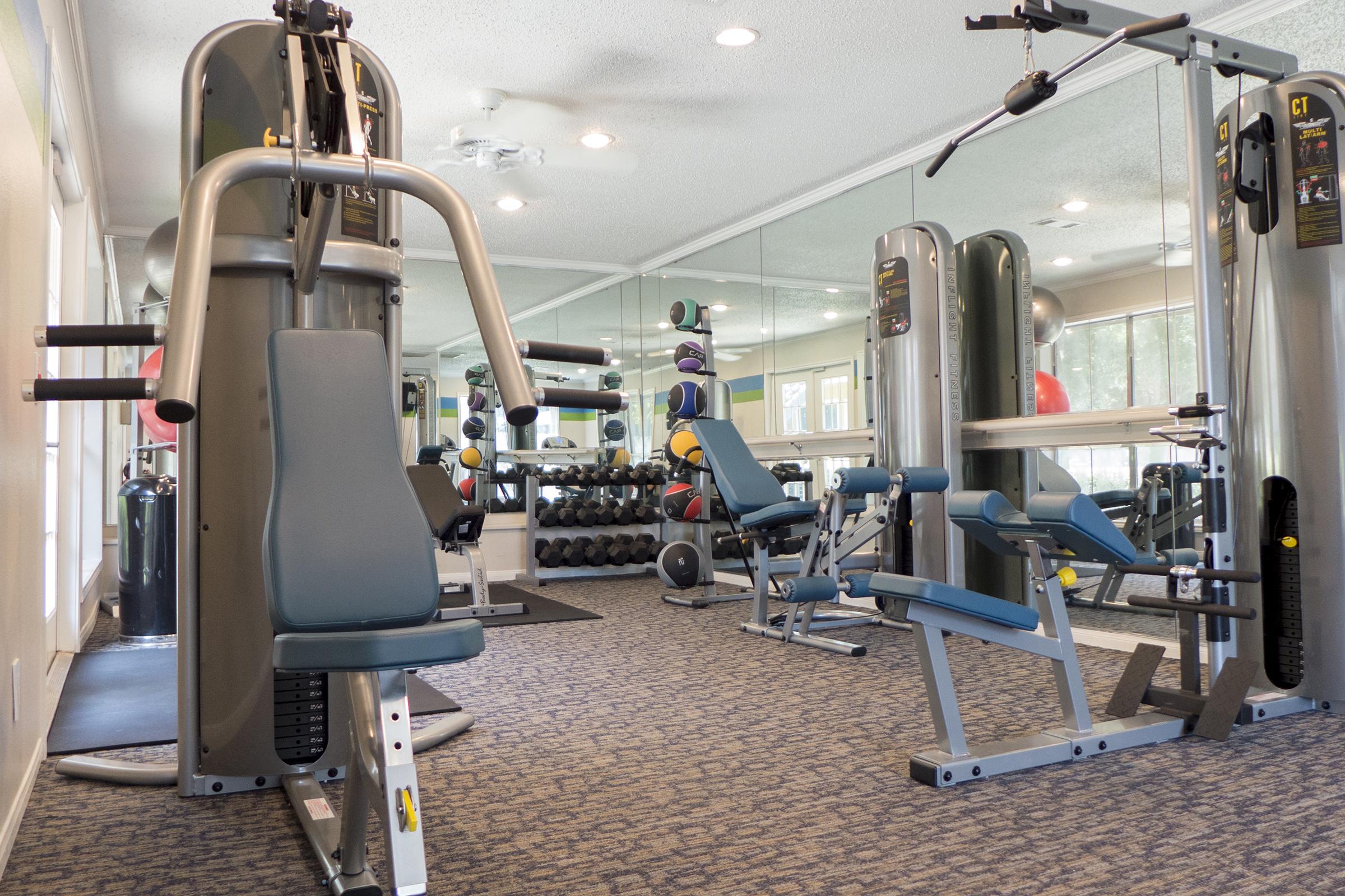 A well-lit gym interior featuring various exercise machines, including a multi-station weight machine, seated row machine, and free weights arranged on racks. The walls are mirrored, enhancing the sense of space. Exercise balls and other fitness equipment are visible, creating a functional workout environment.