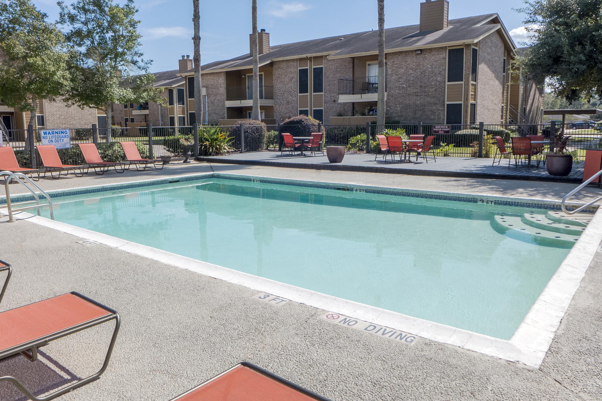 A clear swimming pool surrounded by lounge chairs and a landscaped area. The pool area features a no diving sign, with apartment buildings visible in the background. Trees and chairs add to the relaxed atmosphere of the outdoor space.