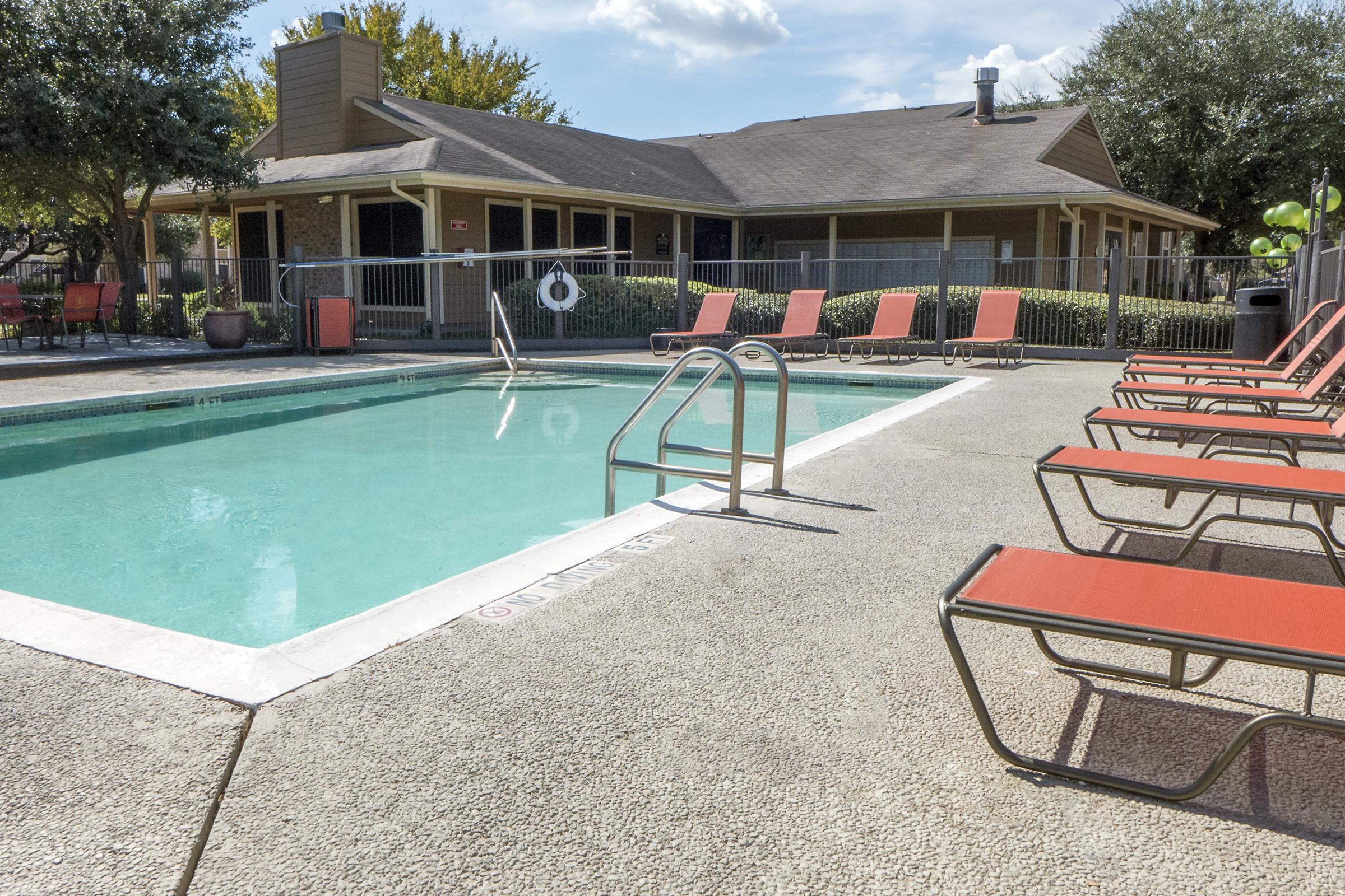 A clear swimming pool with a ladder, surrounded by orange lounge chairs. In the background, a single-story building with large windows and a fenced area. Lush greenery and trees are visible around the pool area, under a bright blue sky with a few clouds.