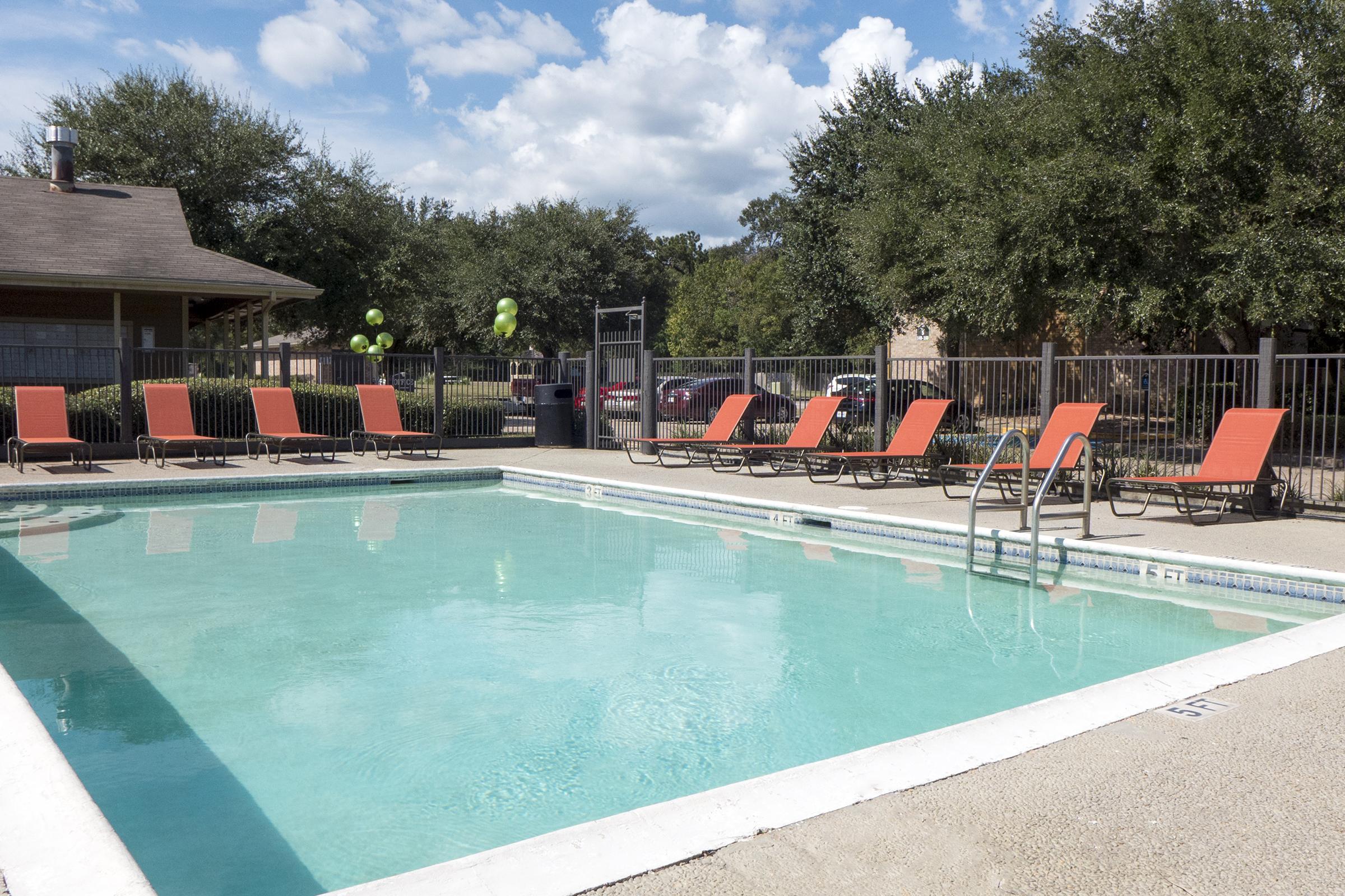 A clear swimming pool surrounded by lounge chairs on a sunny day. There are green trees in the background and a building with large windows. A pool ladder leads into the water, and the area is enclosed by a metal fence.