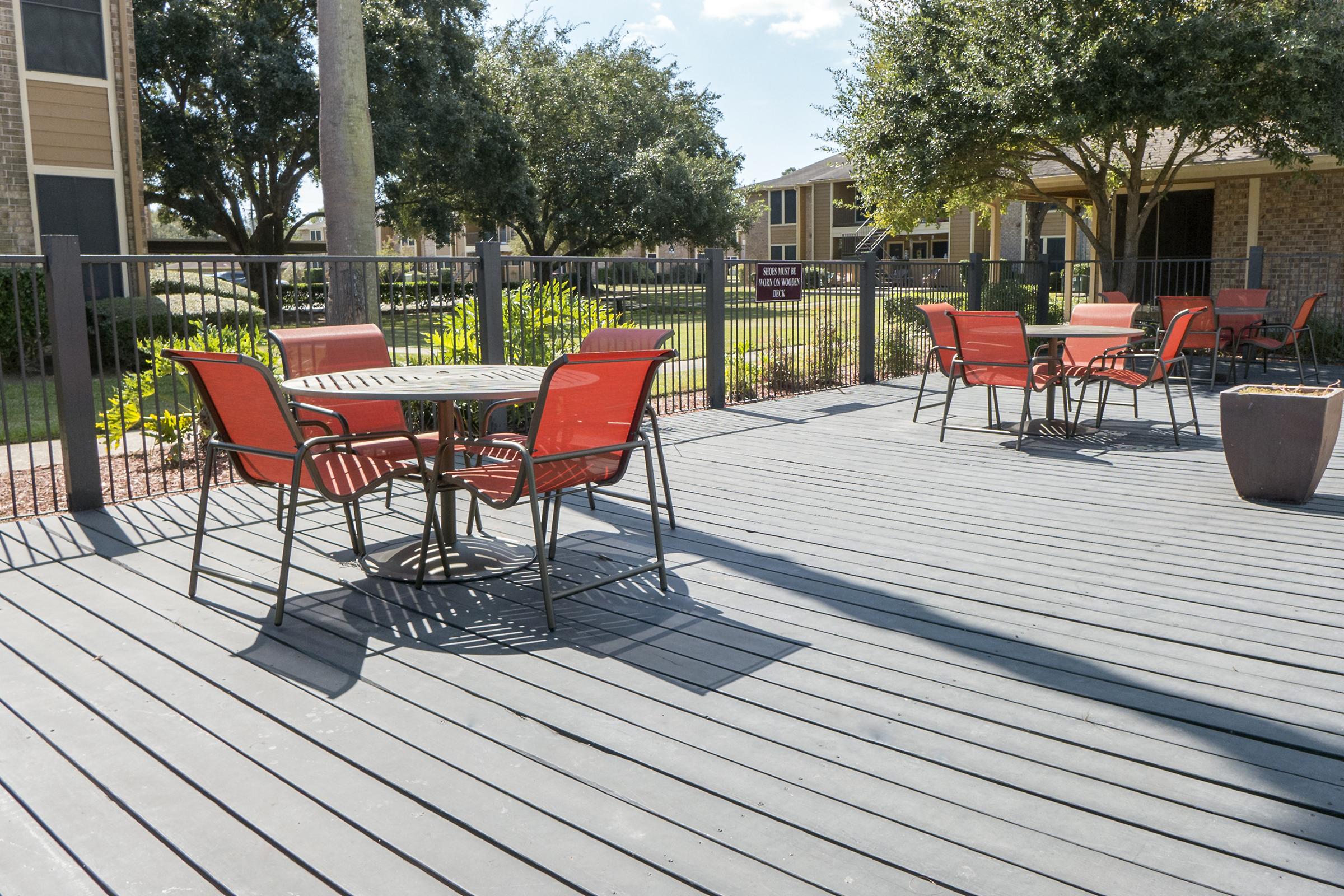 Outdoor patio area with a wooden deck, featuring a round table surrounded by four red metal chairs. In the background, there are green trees and apartment buildings, with a sunny sky above. A plant pot is positioned next to the table, adding to the inviting atmosphere.