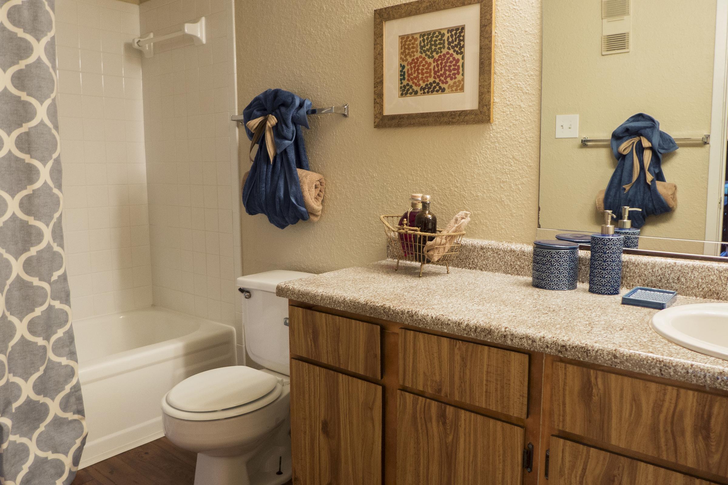 A clean bathroom featuring a beige wall, a shower with a white curtain, a bathtub, and a wooden vanity. The vanity is topped with a sink, decorative items, and toiletries, while a framed artwork hangs on the wall above. Blue accents add color to the space.