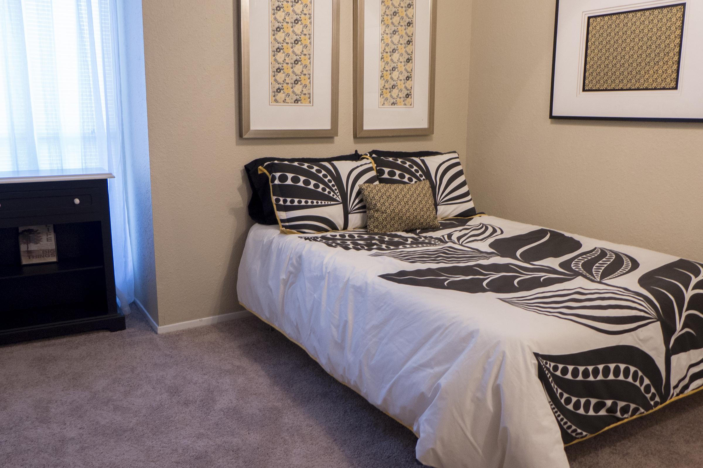 A cozy bedroom featuring a double bed with a black and white leaf-patterned duvet and two decorative pillows. The walls are light-colored, adorned with framed artwork. A small bedside table holds books, and sunlight streams through sheer curtains, creating a warm, inviting atmosphere.