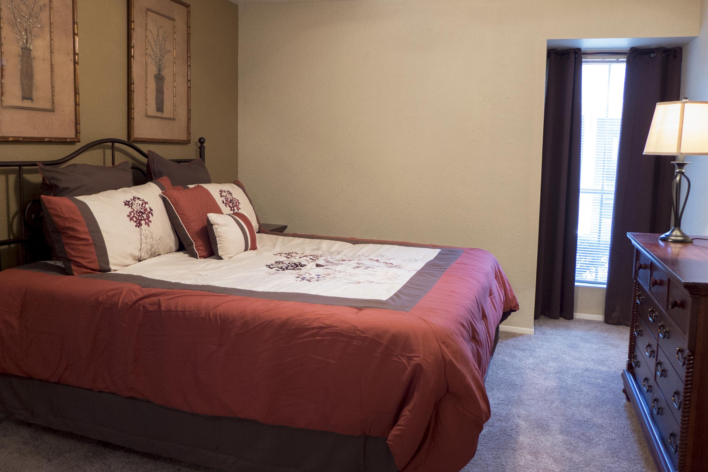 A cozy bedroom featuring a queen-sized bed with a burgundy and cream comforter, accented by decorative pillows. Two framed artworks hang above the bed, and a wooden dresser is positioned nearby. A window with light curtains allows natural light to enter, and a bedside lamp provides additional illumination.
