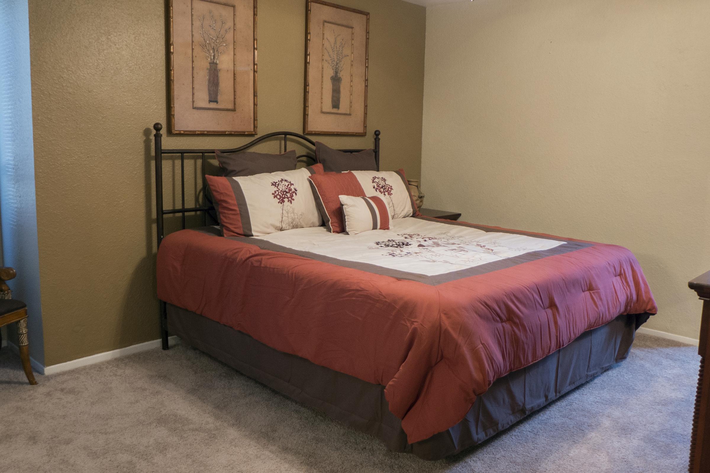 A cozy bedroom featuring a neatly made bed with maroon and cream bedding, adorned with decorative pillows. The walls are painted a soft beige, and two framed art pieces hang above the bed. A bedside table and a chair are visible, adding to the inviting atmosphere of the room.