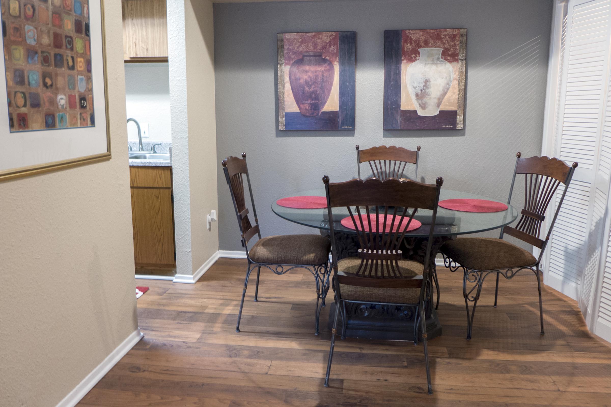 A cozy dining area featuring a round glass table with a decorative metal base and four wooden chairs, two of which have cushions. The walls are adorned with two abstract art pieces depicting vases. Hardwood flooring complements the inviting atmosphere, with a glimpse of a kitchen in the background.