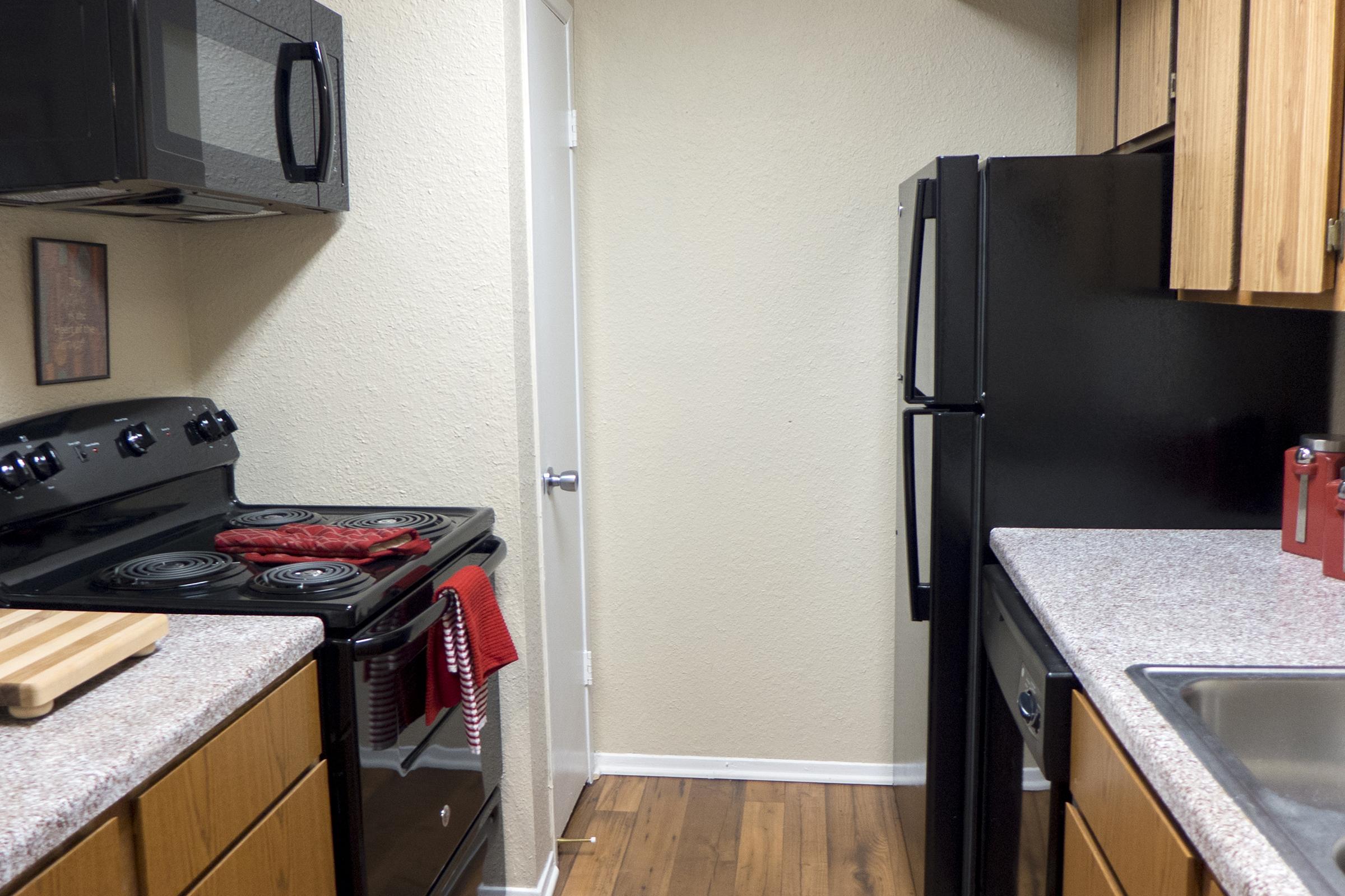 A small kitchen featuring a black stove, microwave, and refrigerator. The countertops are light-colored with a cutting board and a red towel on the stove. Wooden cabinets line the walls, and there is a door leading out of the kitchen. The floor is composed of brown wood-like planks.