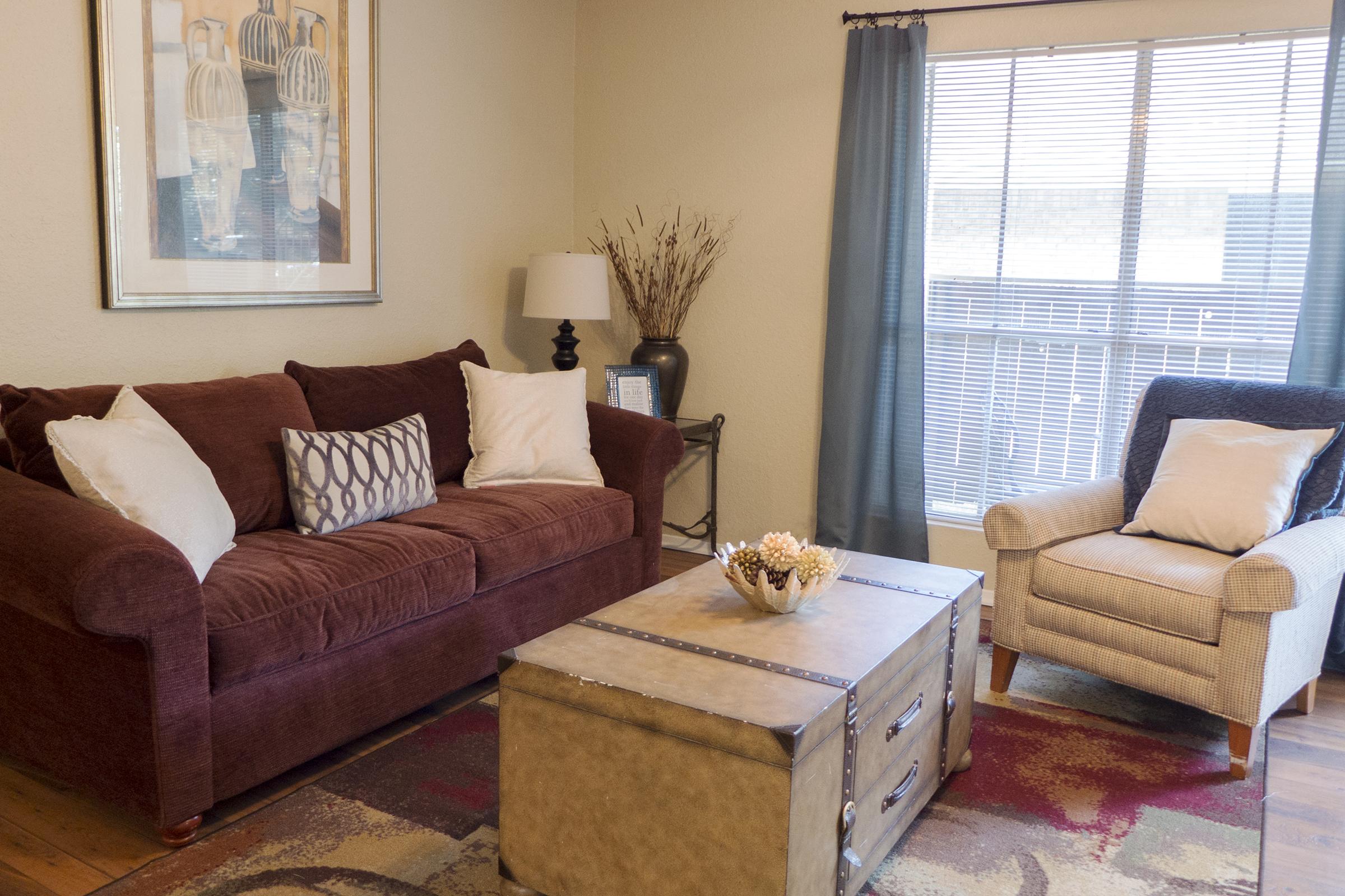 A cozy living room with a burgundy sofa adorned with decorative pillows, a light-colored armchair, and a metal coffee table centered on a patterned rug. Natural light streams in through a large window with blue curtains, adding warmth to the inviting space decorated with a framed artwork and a lamp.