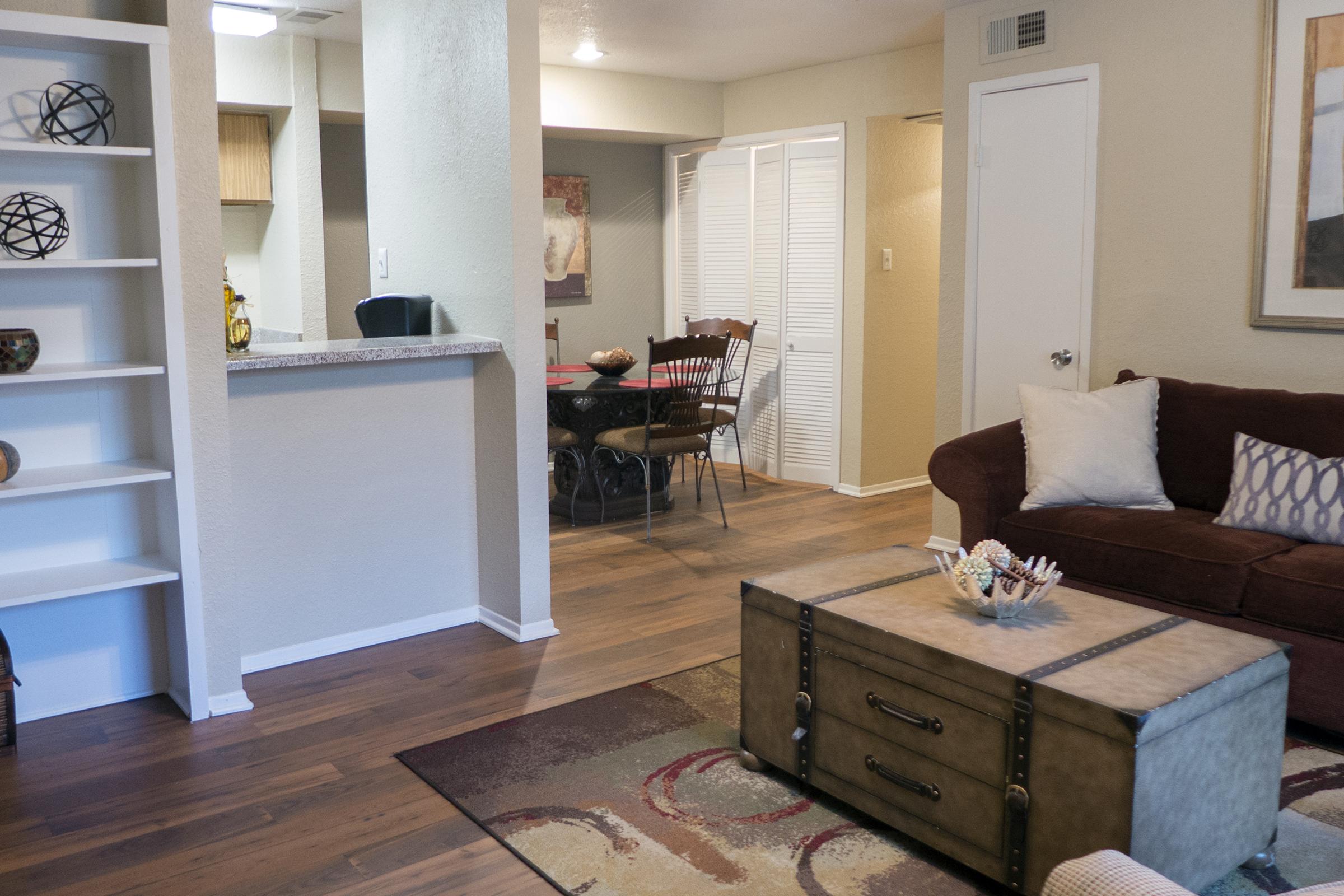 Living room and dining area of a modern apartment. The space features a brown sofa, a decorative coffee table, and a patterned rug. There is a bookshelf with decorative items, a dining table with chairs in the background, and light-colored walls. The overall aesthetic is cozy and welcoming.