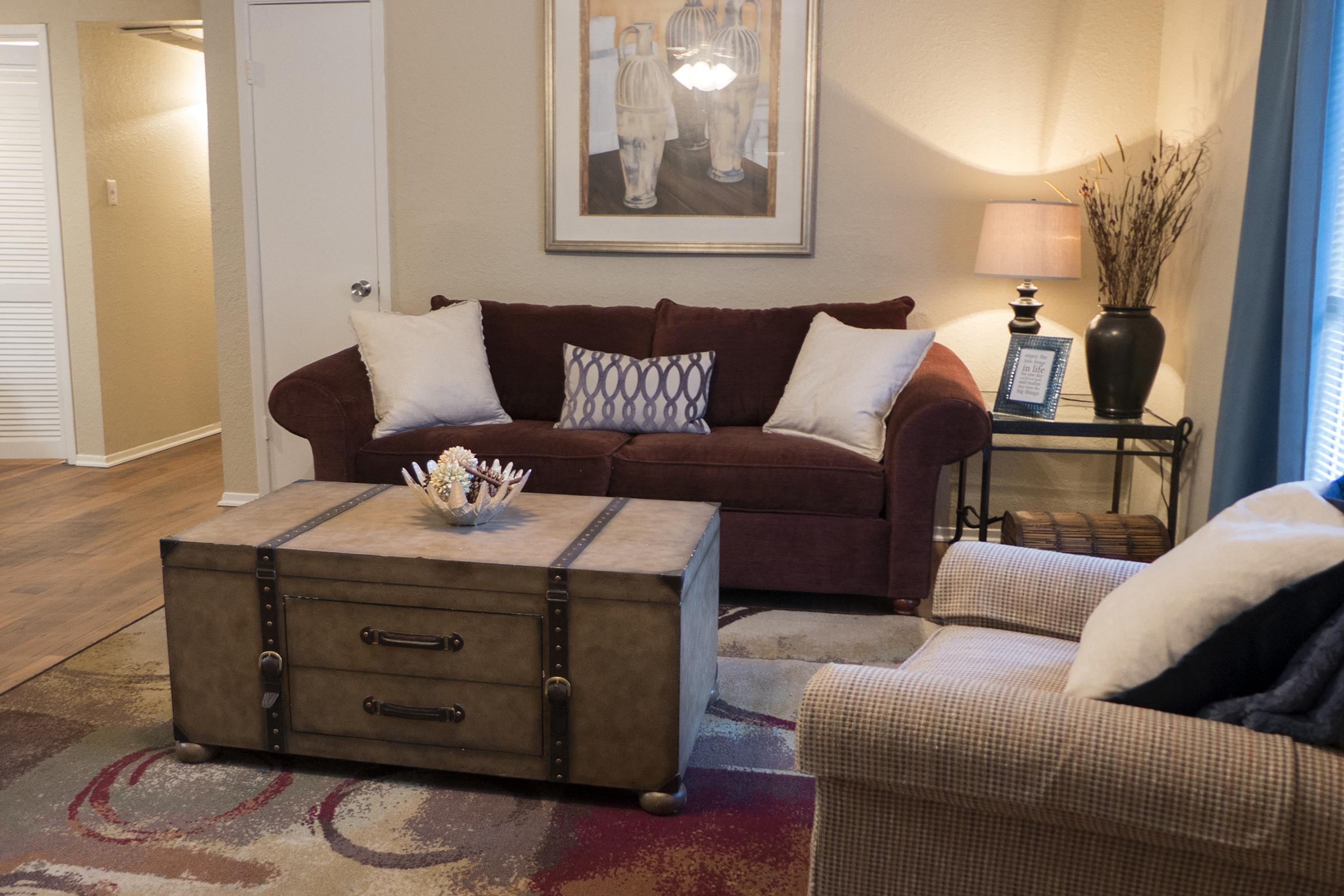A cozy living room featuring a burgundy sofa with white and patterned cushions, a rustic coffee table with metal accents, and a light-colored armchair. Decorative items include a lamp, framed artwork on the wall, and a vase with dried plants. Soft area rug and natural light enhance the inviting atmosphere.