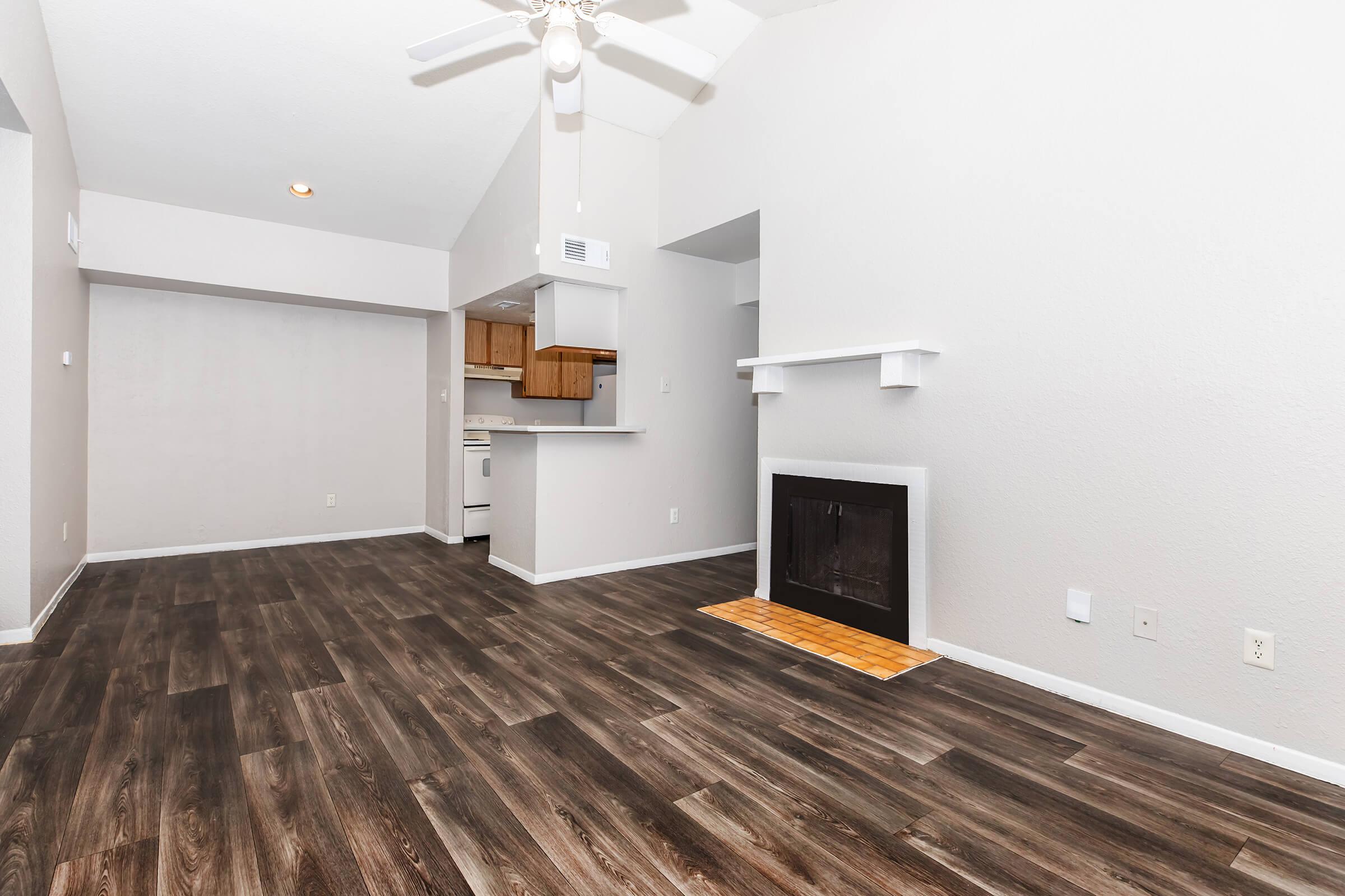 A spacious living area featuring light-colored walls and dark wood-like flooring. There's a ceiling fan overhead, a small fireplace with a tiled hearth, and an open view leading to a kitchen area with wooden cabinets. Natural light fills the room, creating a bright and inviting atmosphere.