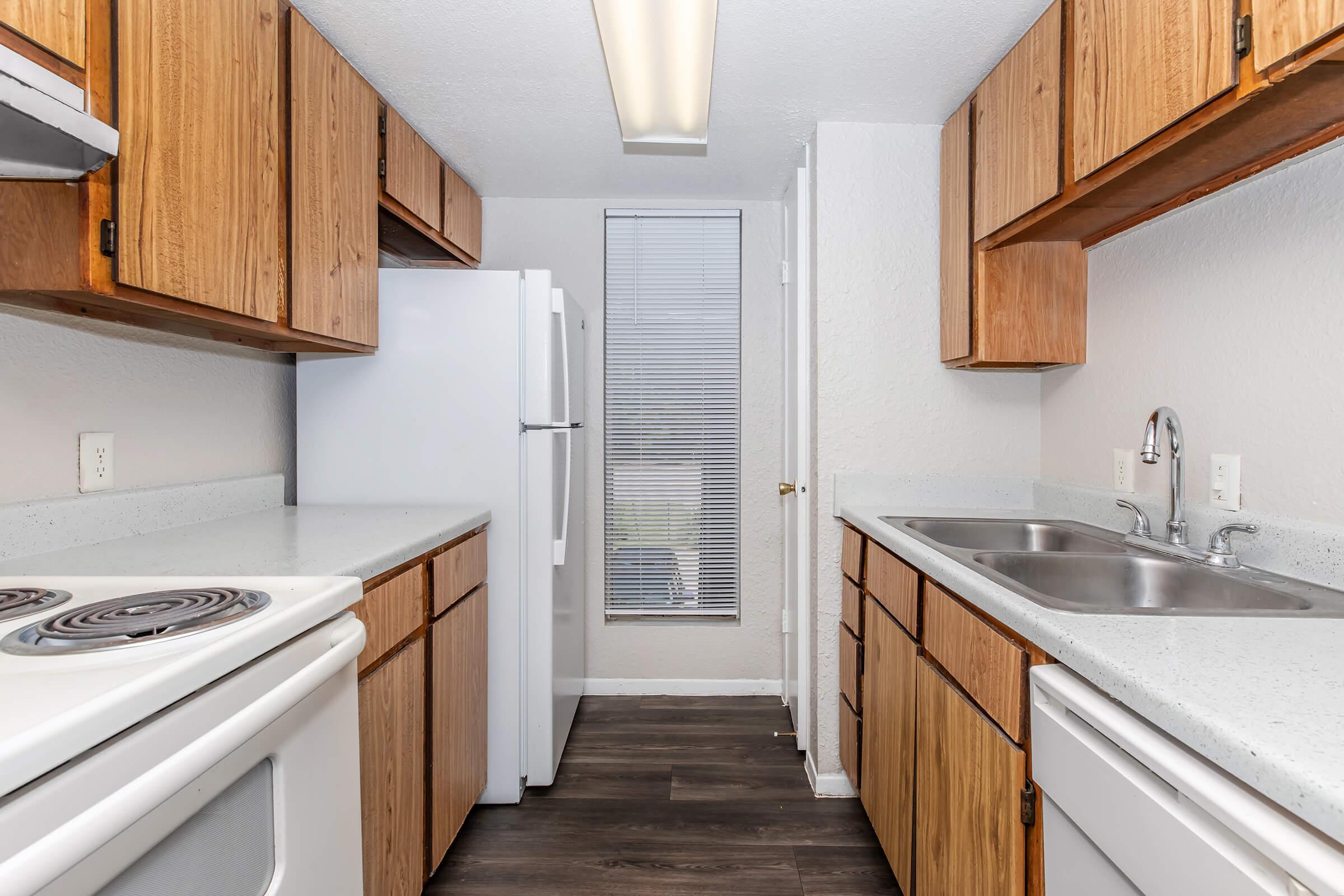 A modern kitchen featuring wooden cabinets, a white refrigerator, a double sink, and a stove. The countertop is light-colored, and there is a window with blinds providing natural light. The flooring is dark, creating a contrast with the cabinetry.