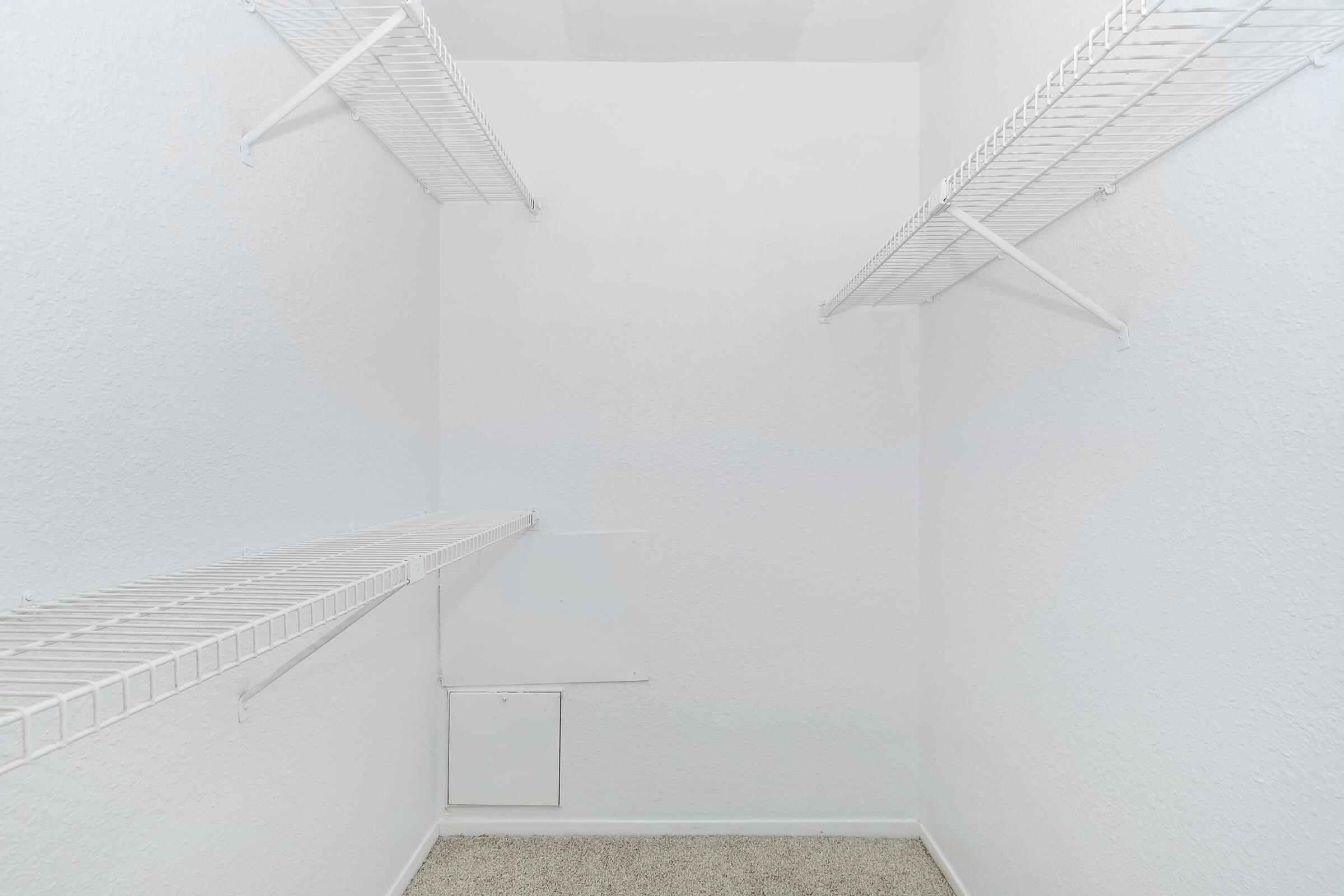 Empty closet with white walls and two wire shelves on opposite sides. The floor is carpeted in a light color, and a small access panel is visible on one wall. The overall appearance is clean and minimalist.