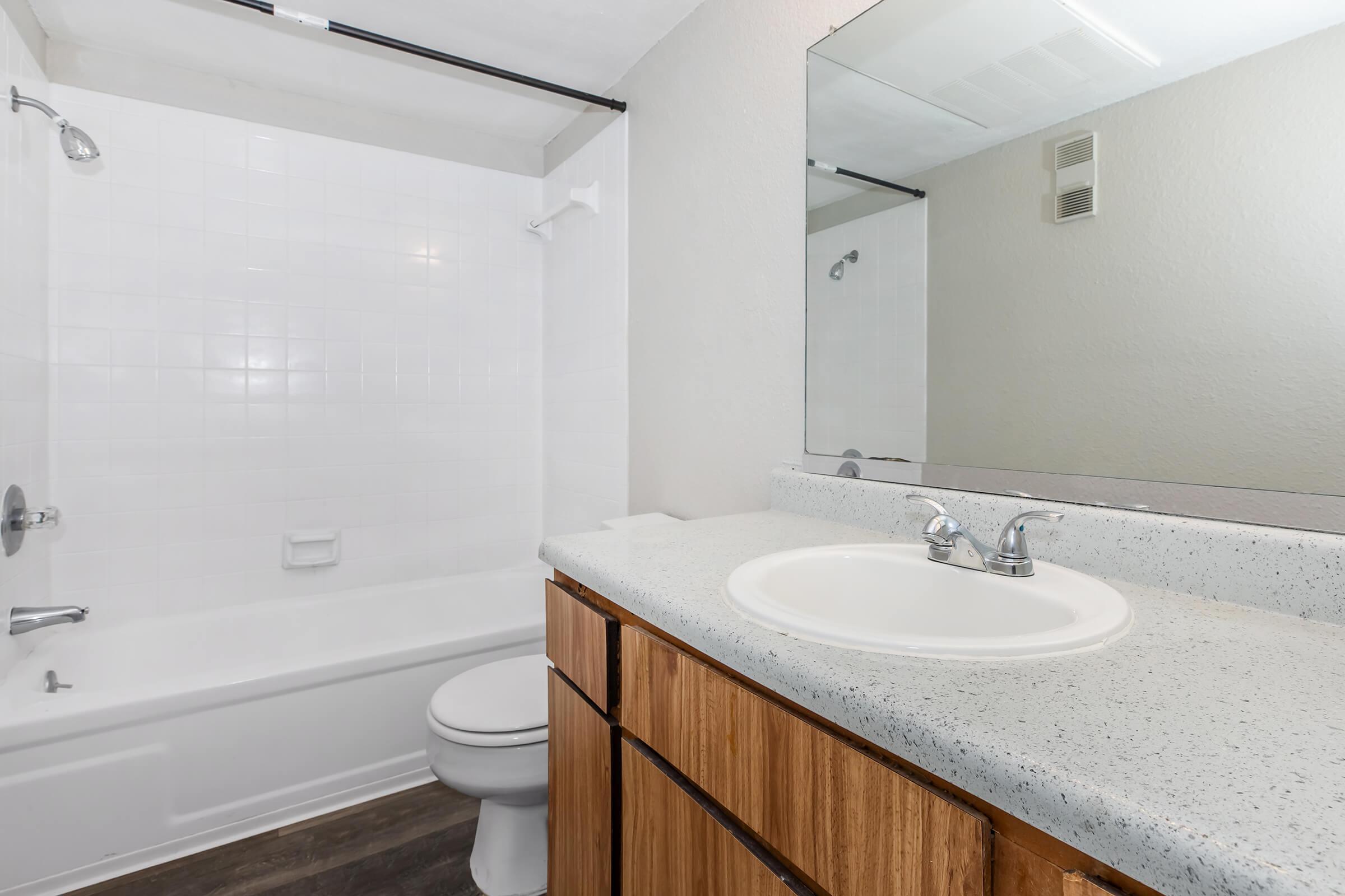 A clean and modern bathroom featuring a bathtub with a showerhead, a white tiled wall, a circular sink with a speckled countertop, and wooden cabinets. A large mirror reflects the space, and the flooring has a dark wood appearance, creating a contemporary look.
