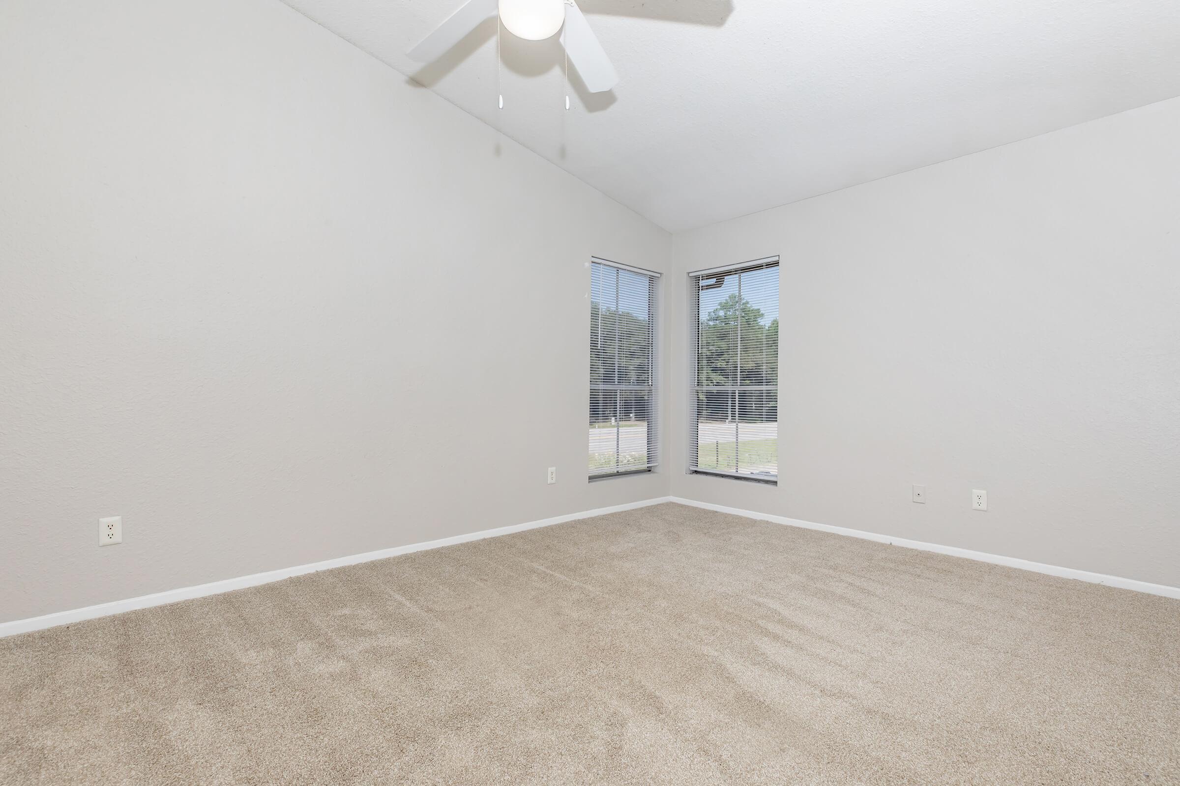 A spacious, empty room with beige carpet and neutral gray walls. There are two windows allowing natural light, and a ceiling fan is mounted on the ceiling. The room has a minimalist look with a clean and uncluttered appearance, perfect for personal customization.