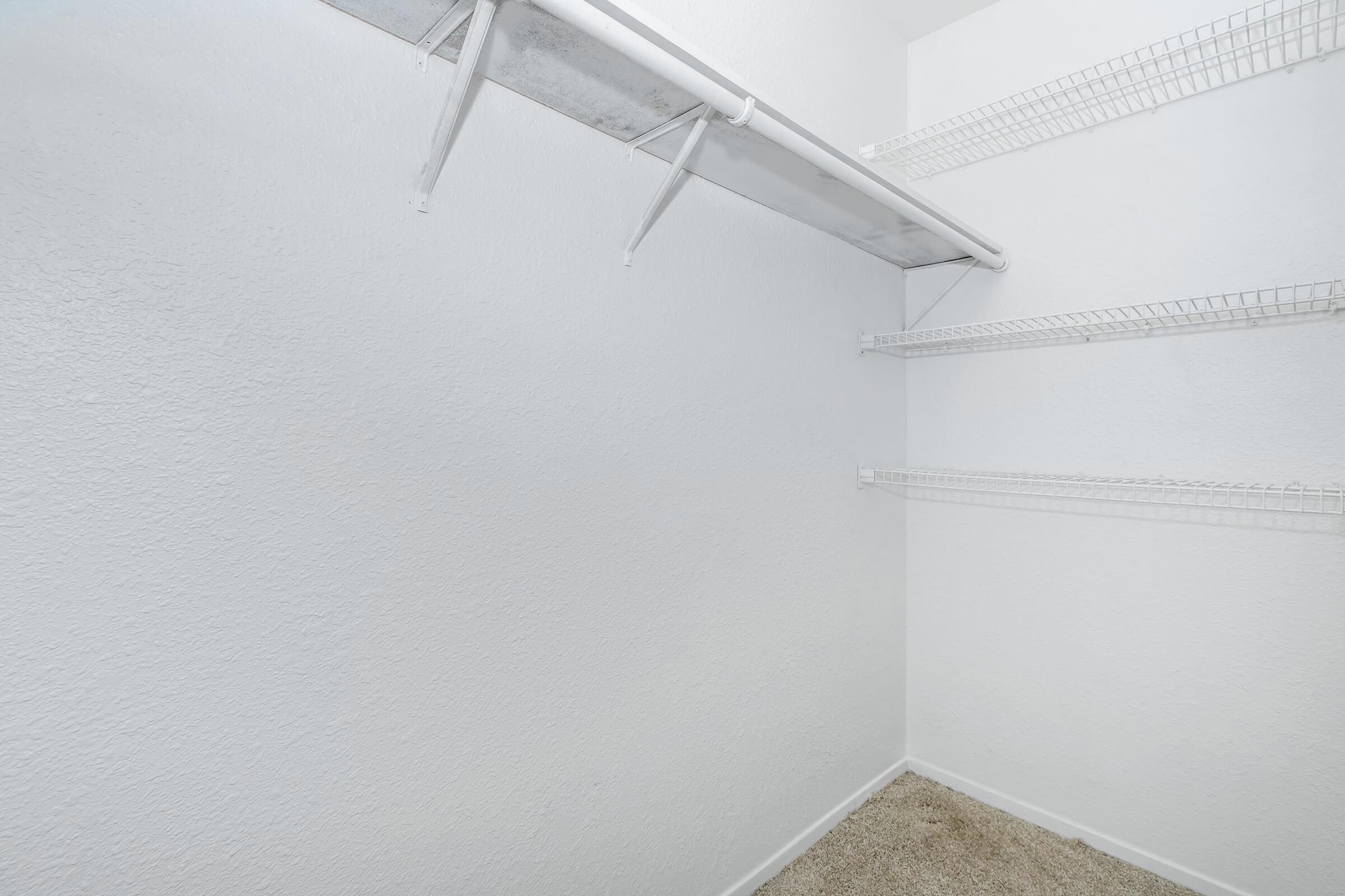 Empty closet with white walls and a single white shelving unit. The floor is covered with a light-colored carpet. The shelving is installed at various heights, providing storage space. The overall appearance is clean and minimalistic.