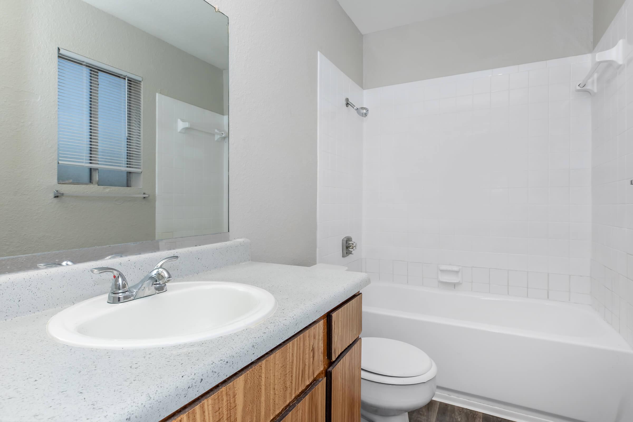 A bathroom featuring a white bathtub and shower combination, a sink with a chrome faucet, and wooden cabinetry. There is a large mirror above the sink and a window with blinds allowing natural light to enter the space. The walls are painted in a light color, creating a bright and clean atmosphere.